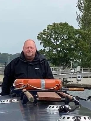 Man in a black jacket standing on a boat with a life preserver and trees in the background.