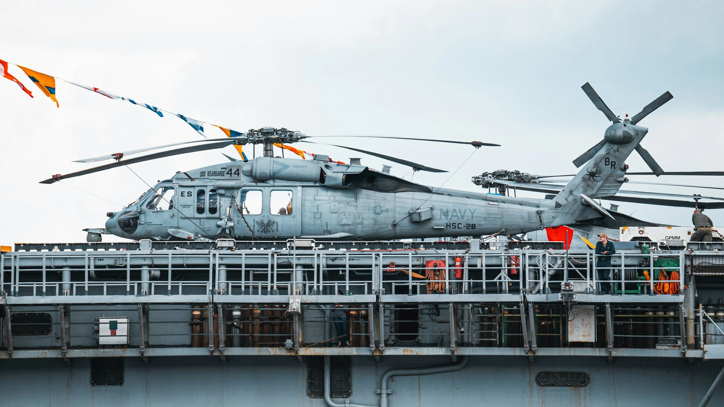 Two military helicopters on a navy aircraft carrier deck, decorated with colorful flags, with crew members attending to them and equipment visible on the deck.