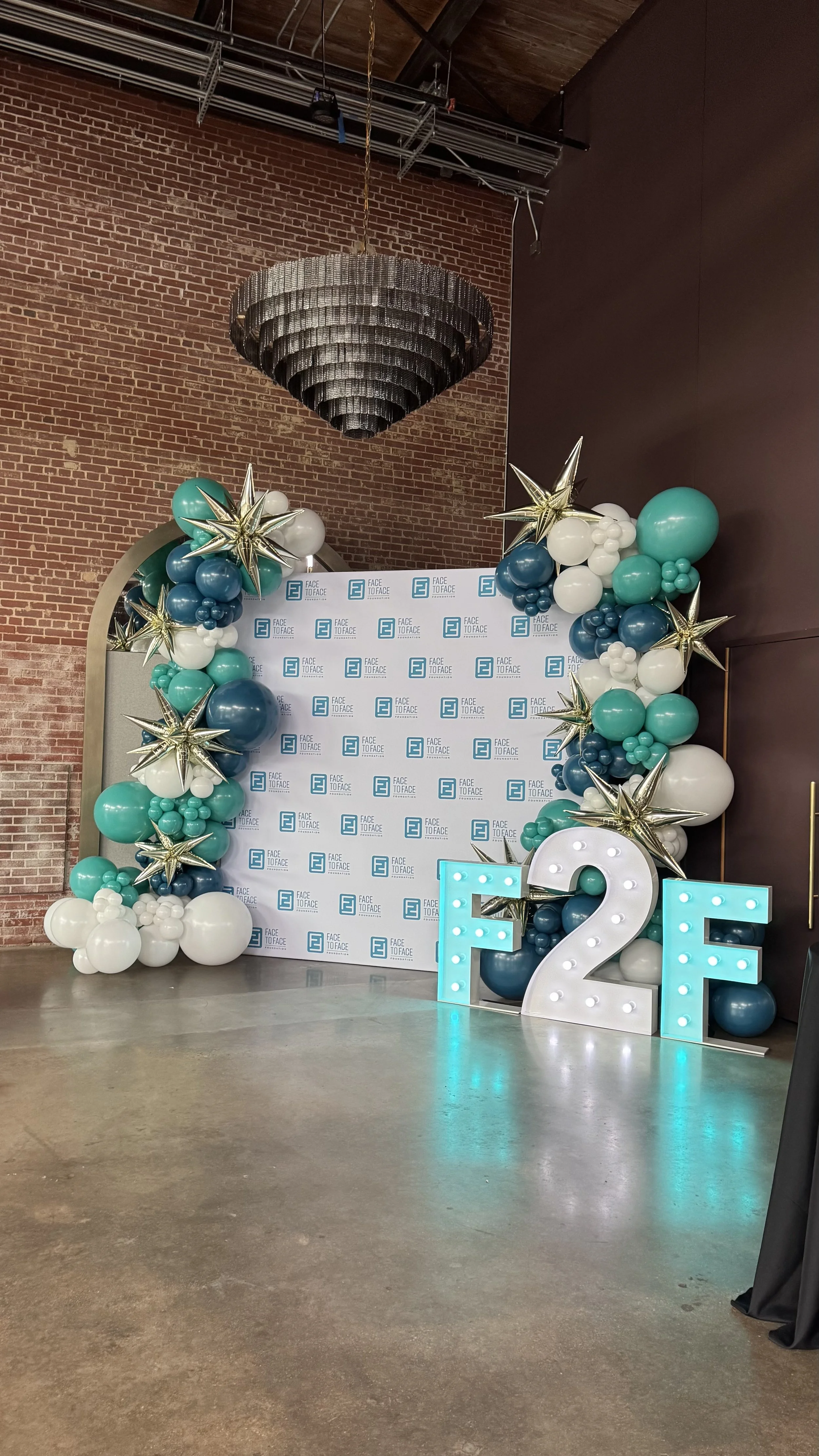 Decorative backdrop with balloons and a large illuminated number two, celebrating a 25th anniversary or milestone, with a brick wall, chandelier, and branded background for an event.