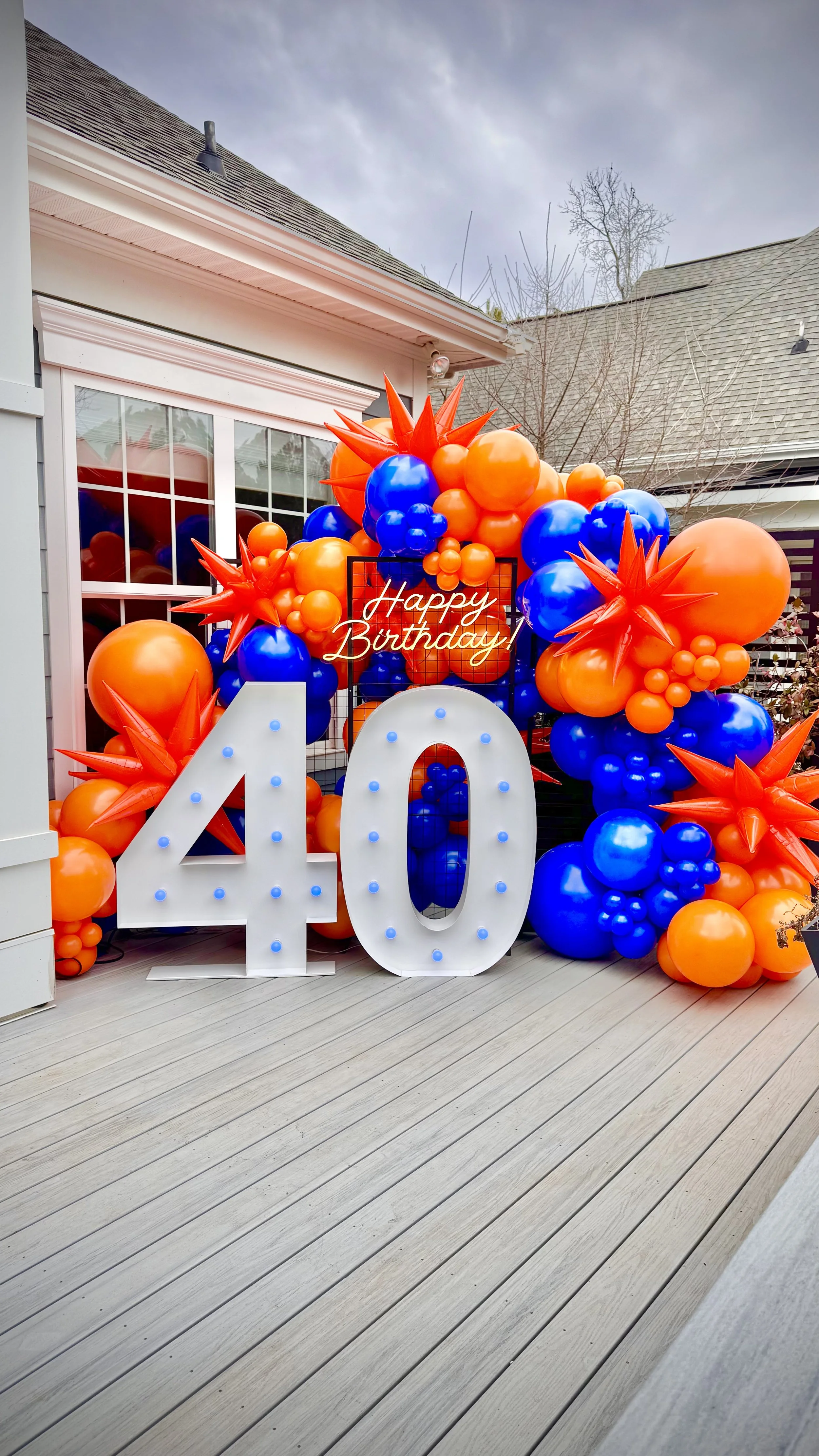 Colorful birthday decoration with orange and blue balloons, a neon sign saying "Happy Birthday," and large illuminated numbers "40" on a wooden deck outside a house.