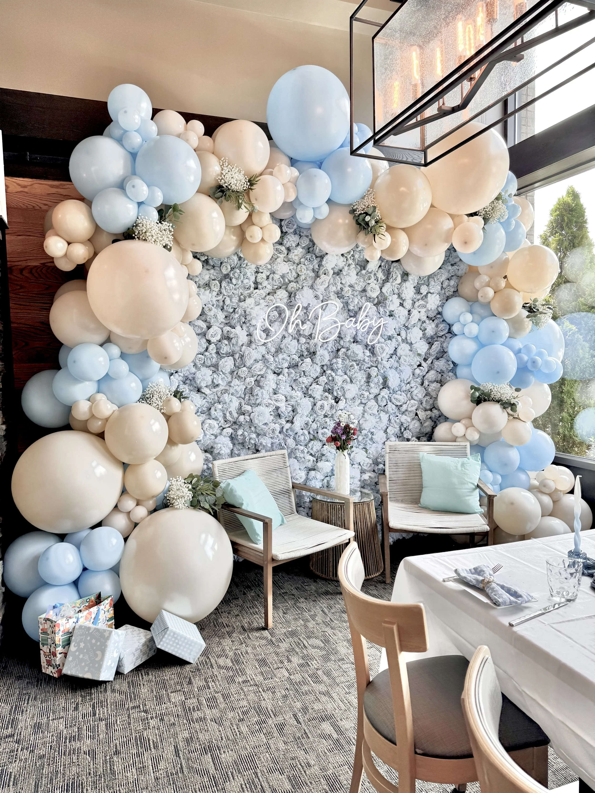 Decorative balloon wall with white and light blue balloons, flowers, and a neon sign that reads 'Oh Baby' in a dining setting with tables, chairs, and a window.