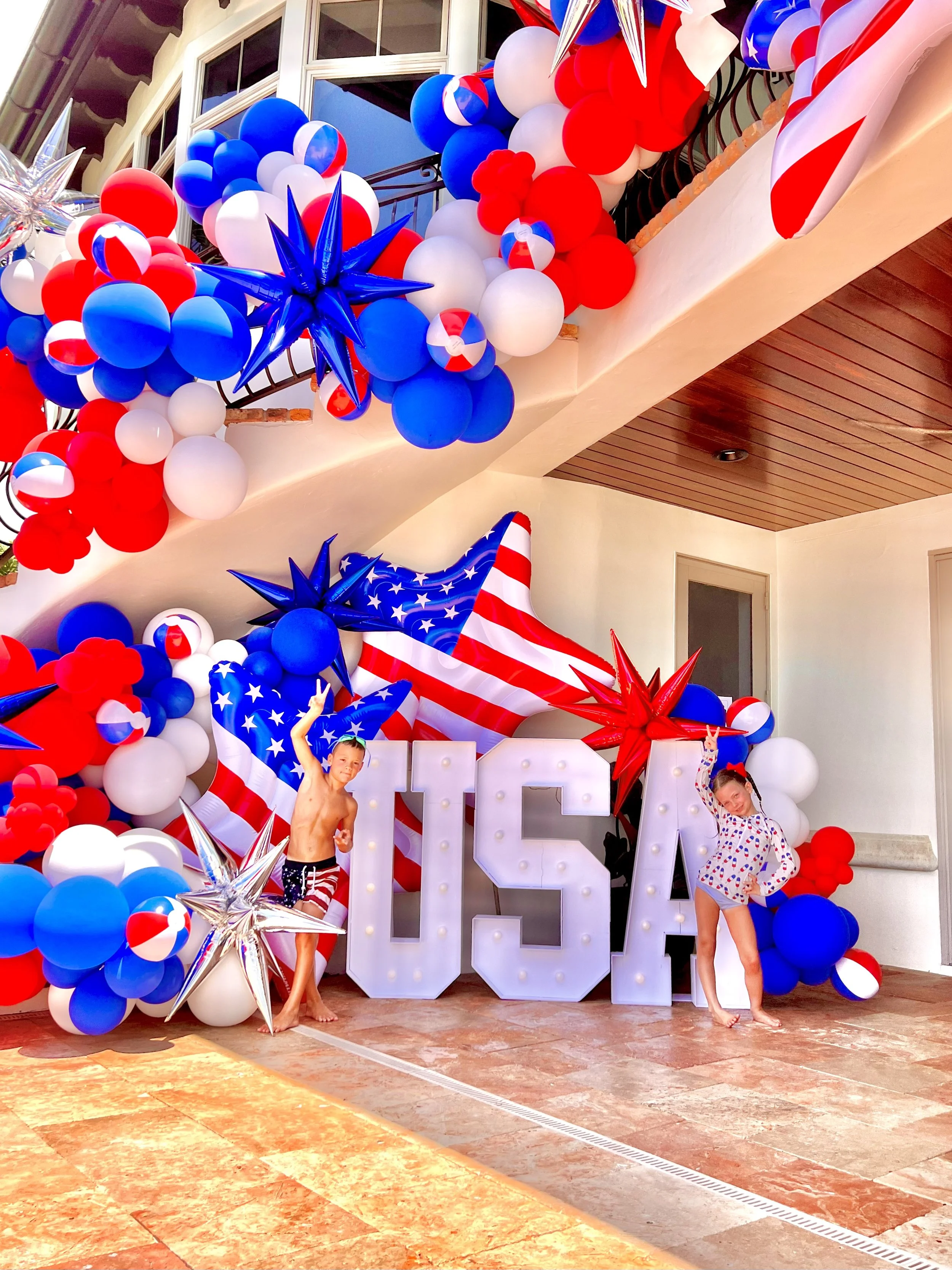 Patriotic Fourth of July celebration decoration with balloons, star-shaped balloons, large lighted letters spelling 'USA', and children in summer clothes at a house porch.