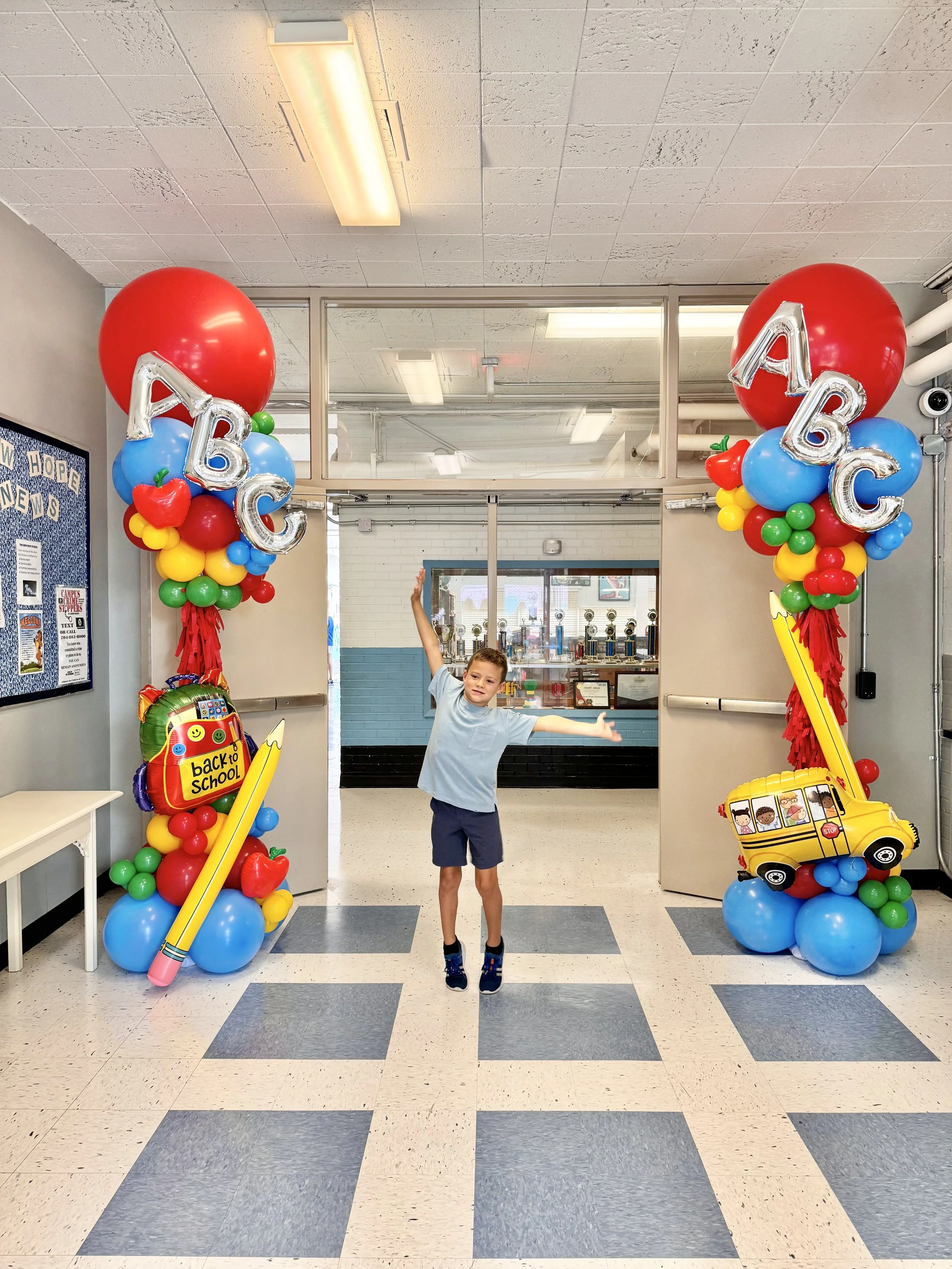 A young boy with outstretched arms jumping in front of decorated school entrance on first day of school, with balloons, 'Back to School' sign, and school-themed decorations.