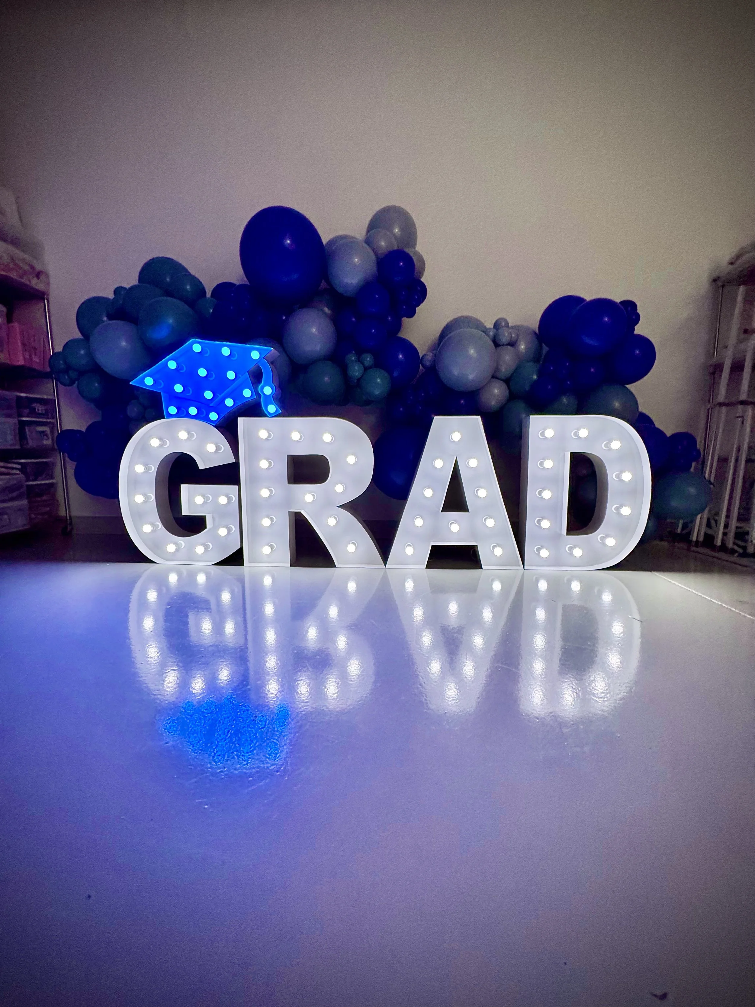 Graduation-themed indoor decoration with illuminated letters spelling 'GRAD' and a blue graduation cap, against a backdrop of blue, gray, and purple balloons.