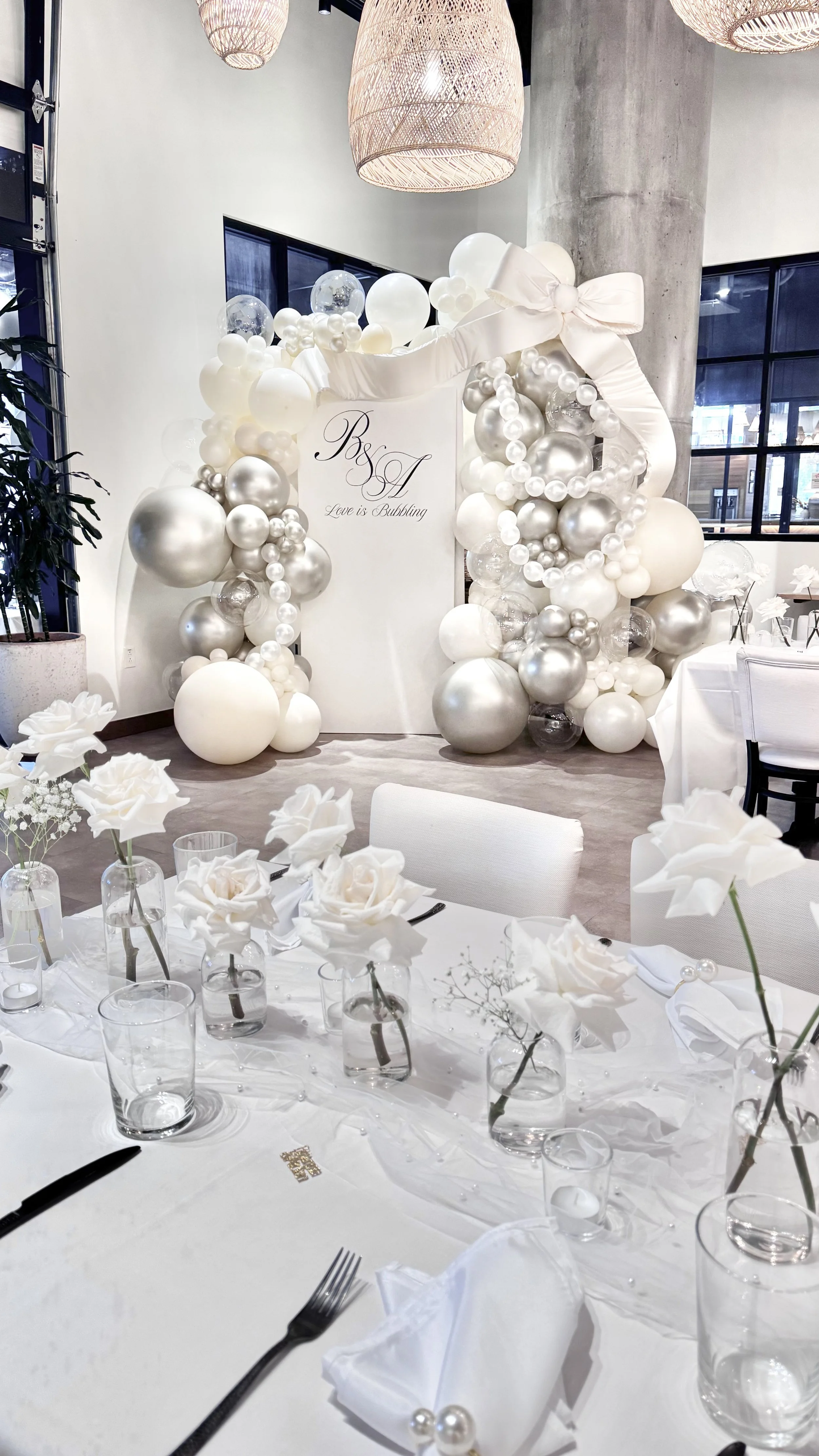 Elegant decorated event space with a balloon arch, white flowers on the table, and a backdrop with 'B & A Love is Bubbling' written on it.