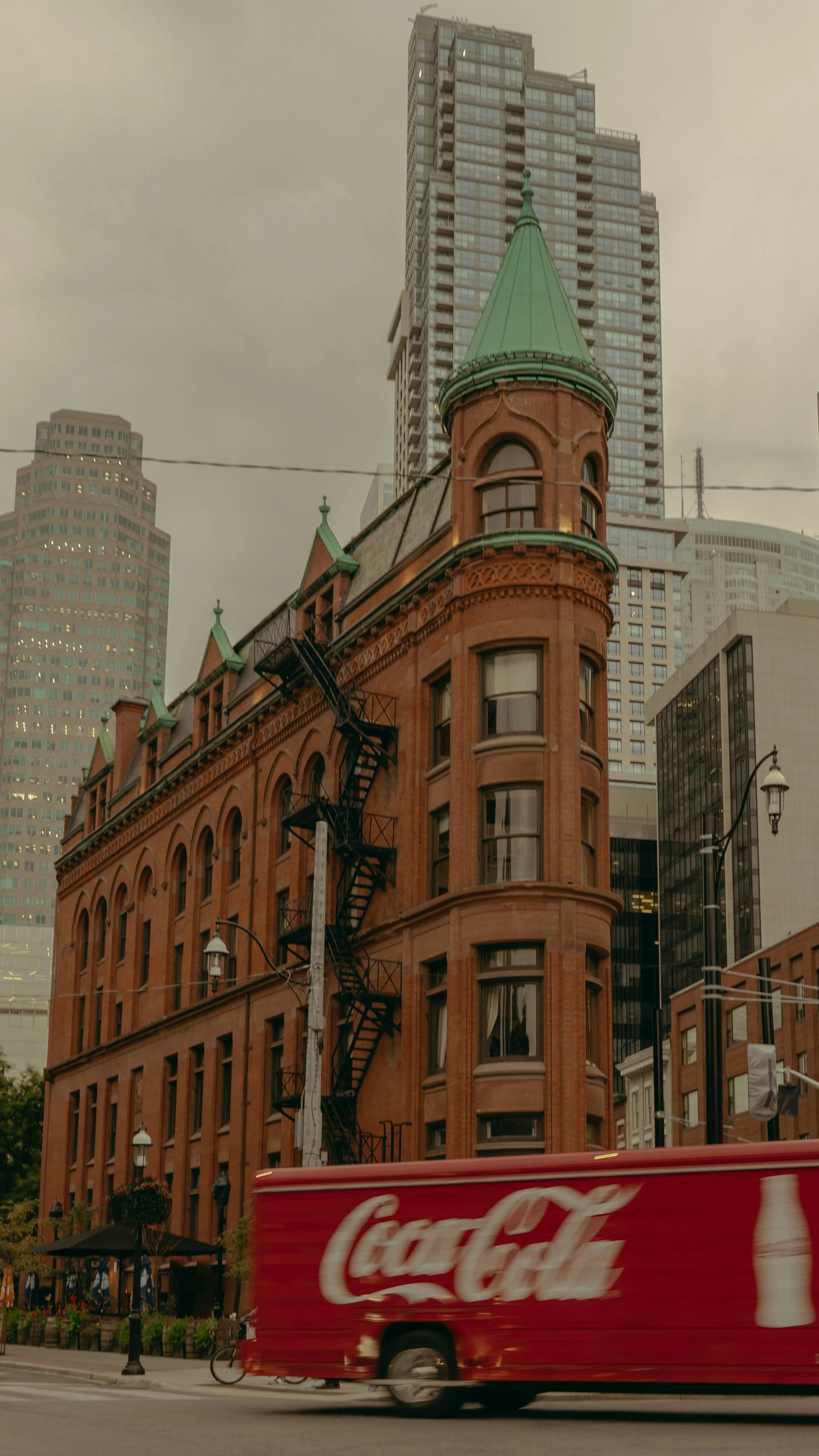 A historic red-brick building with a green steeple against a backdrop of modern skyscrapers, with a Coca-Cola truck passing by in the foreground in an urban city setting.