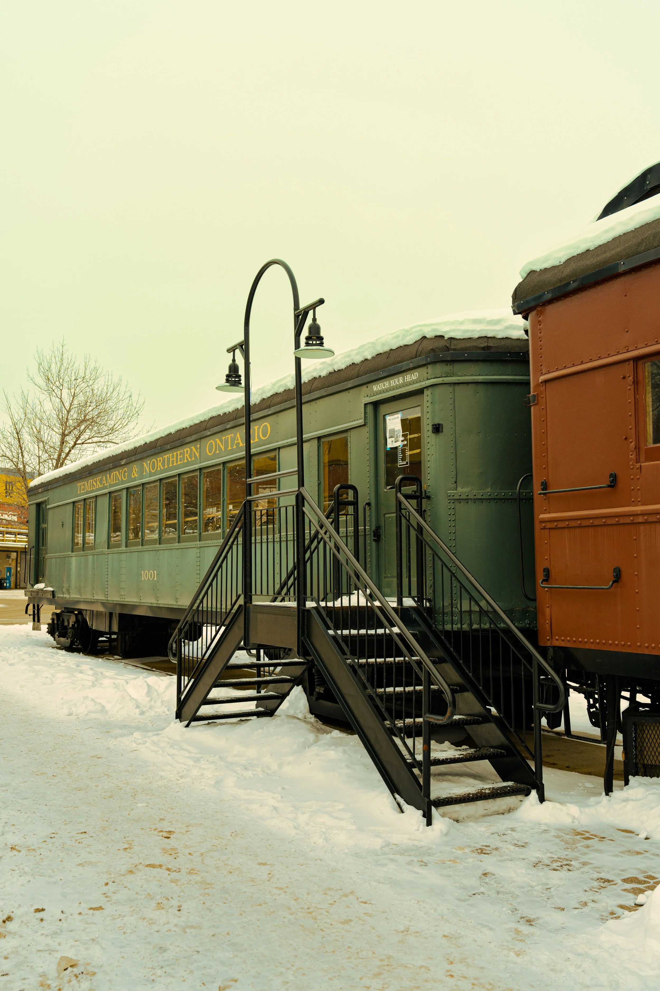 Vintage train car parked on snowy ground with black stairs leading up to its door, and a lamp post nearby. The train car is green with yellow text reading "Temiskaming & Northern Ontario."