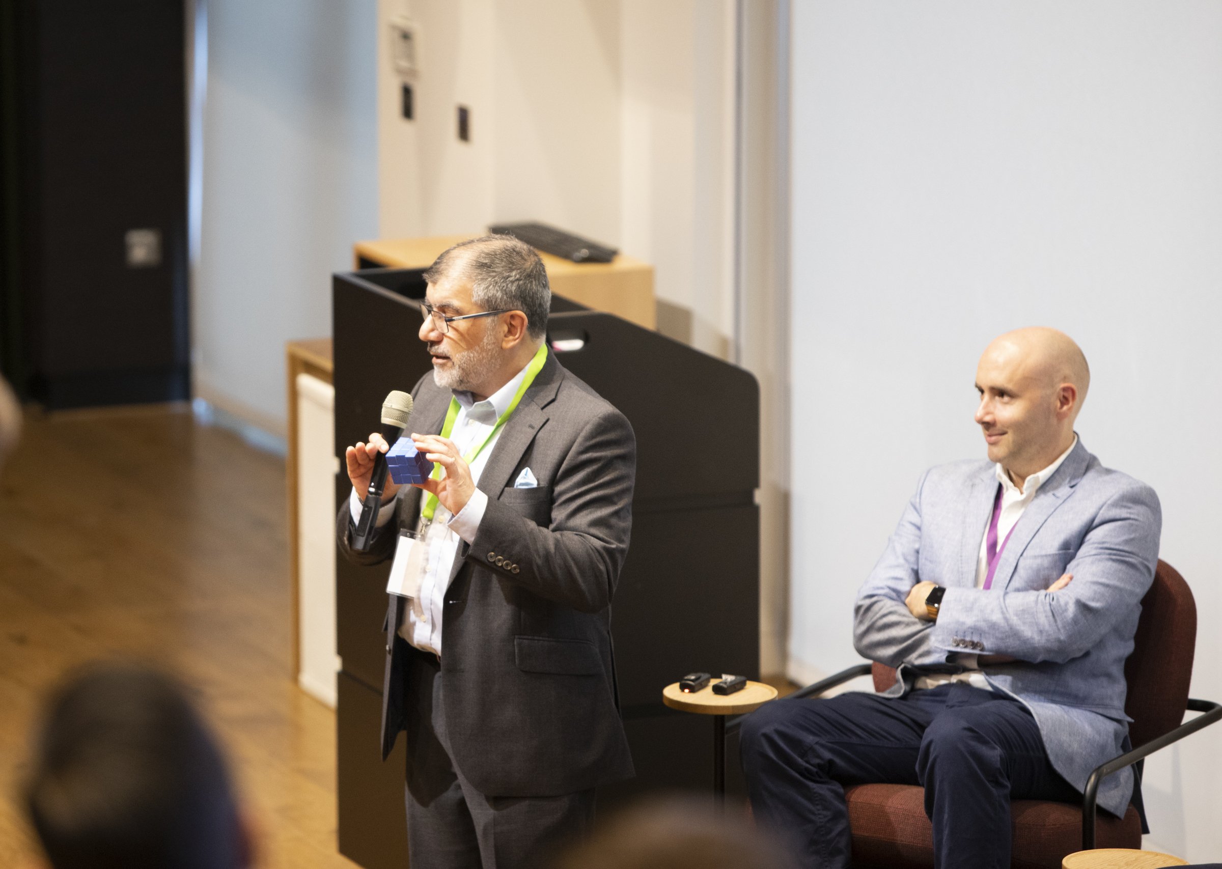 Two men in a conference room, one speaking into a microphone, the other seated.