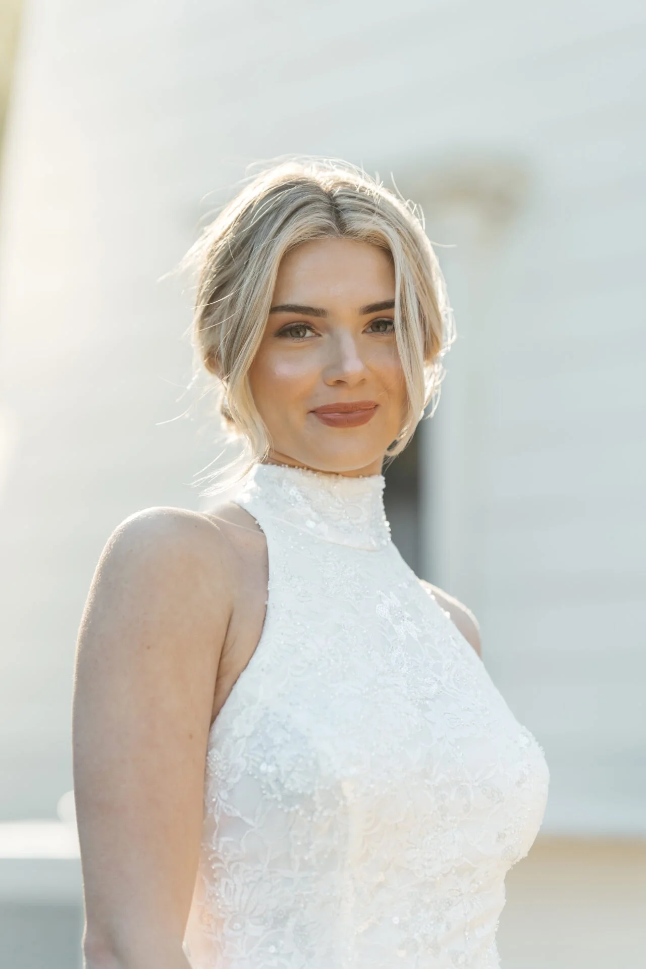 A woman with blonde hair styled in loose waves wearing a white lace sleeveless dress, standing outdoors in natural light.