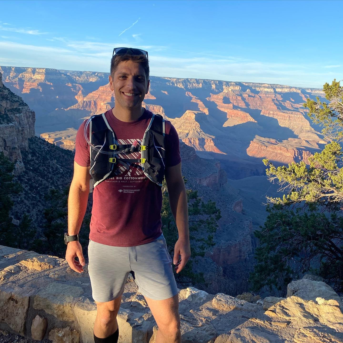 A man standing on a rocky ledge at the Grand Canyon during sunrise or sunset, wearing a maroon T-shirt, light shorts, a black hydration backpack, and sunglasses, smiling with the canyon and blue sky in the background.
