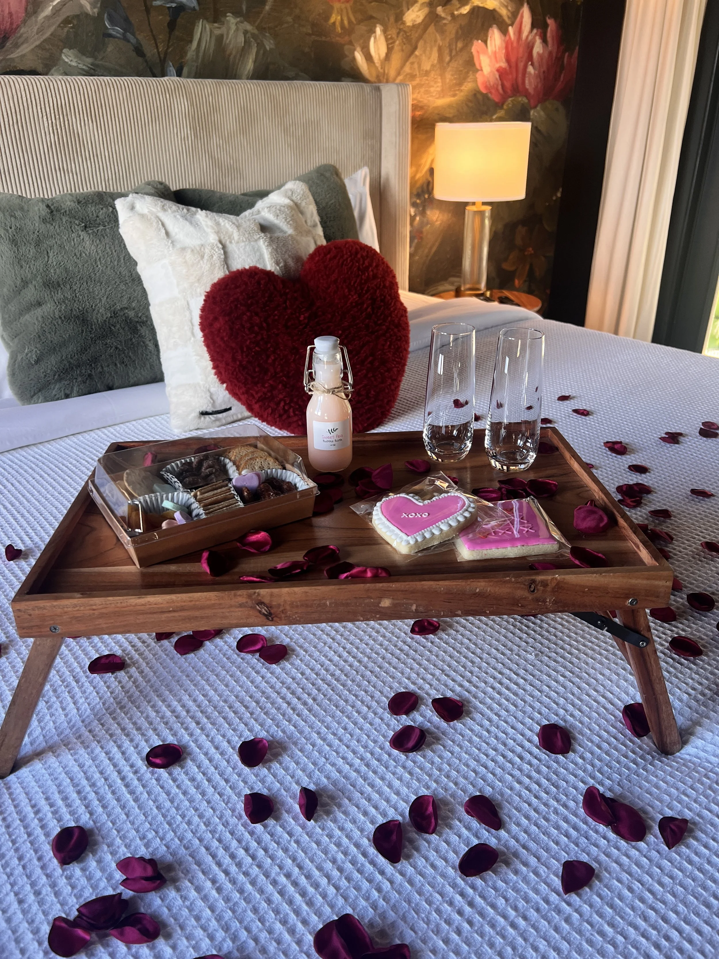 Bed with decorative pillows, rose petals, a wooden tray with sweets, a bottle, and champagne glasses.