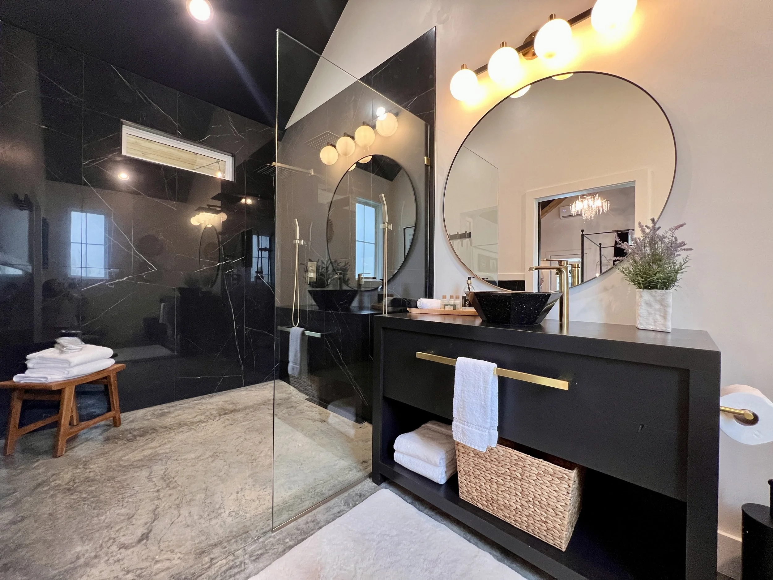 Modern bathroom with a black and gold vanity, round mirror, black marble shower, and decorative elements like folded towels and a potted plant.