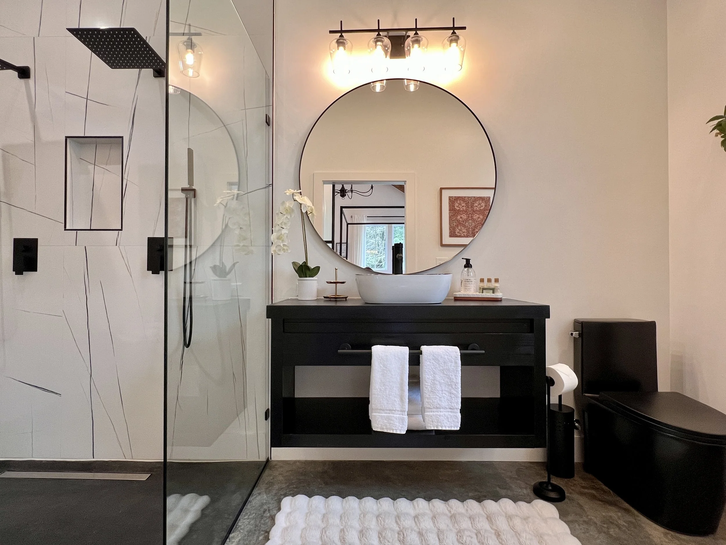 Modern bathroom with glass shower enclosure, black vanity, round mirror, vessel sink, and wall lighting. White towels, decor plants, and toilet visible.
