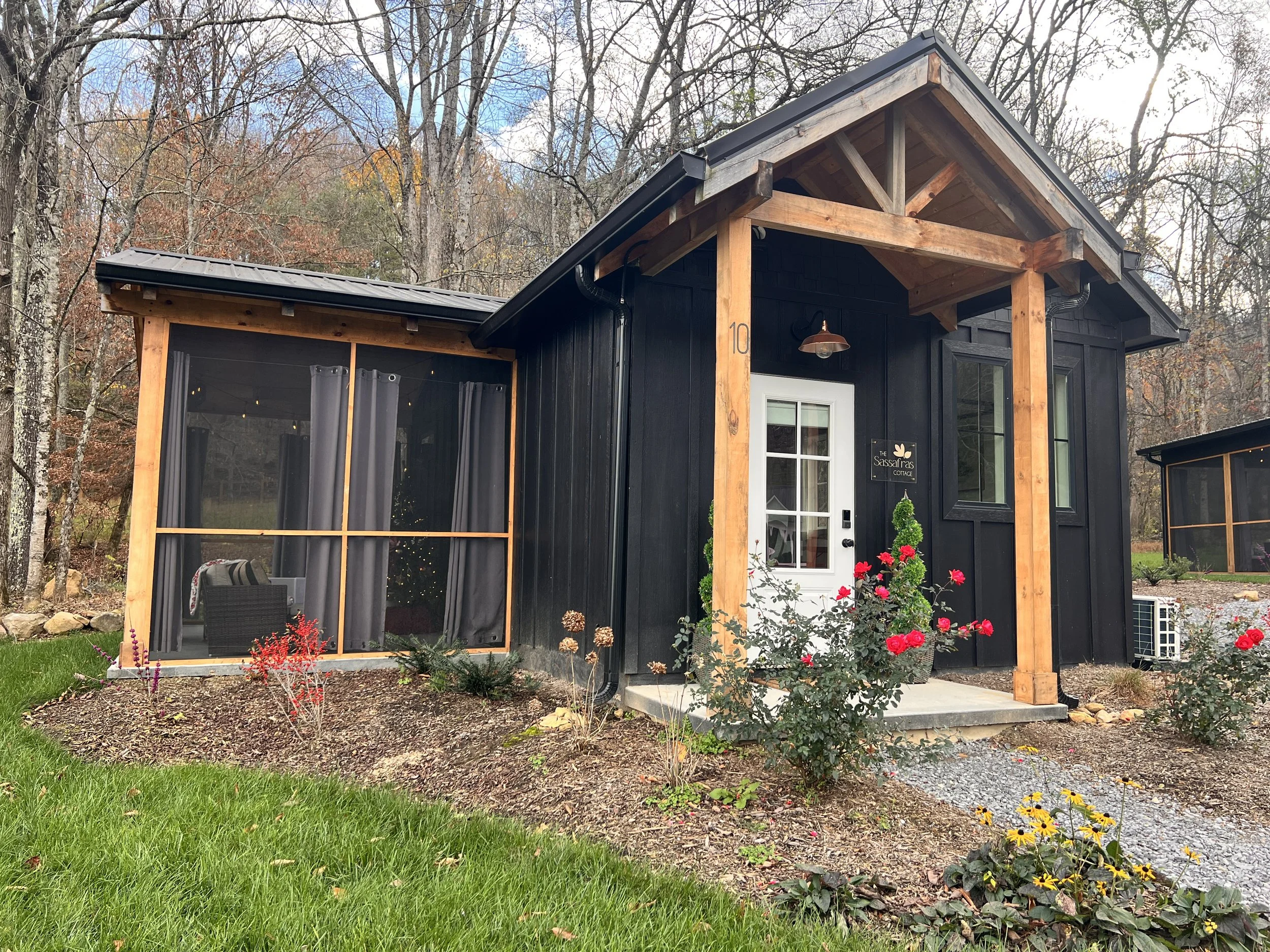 A compact black cabin with wooden trim and a front porch, surrounded by lush greenery and red roses. There's a screened-in outdoor seating area with curtains and a cozy interior visible. The setting is a wooded area during late fall.