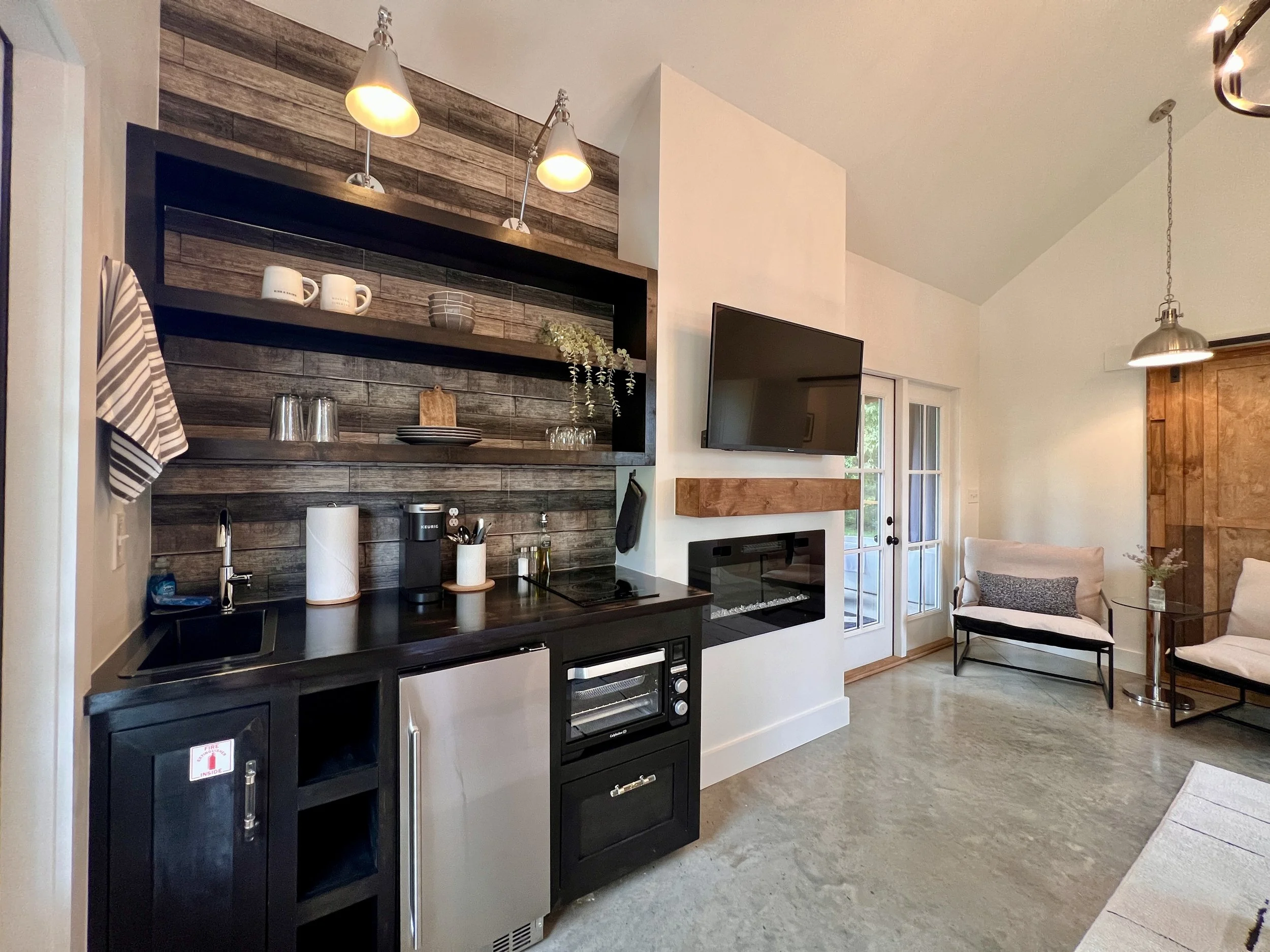 Modern kitchen and living area with dark wood cabinetry, open shelving, mini refrigerator, coffee maker, and wall-mounted TV. Contains a small fireplace below the TV, a French door leading outside, and contemporary seating.
