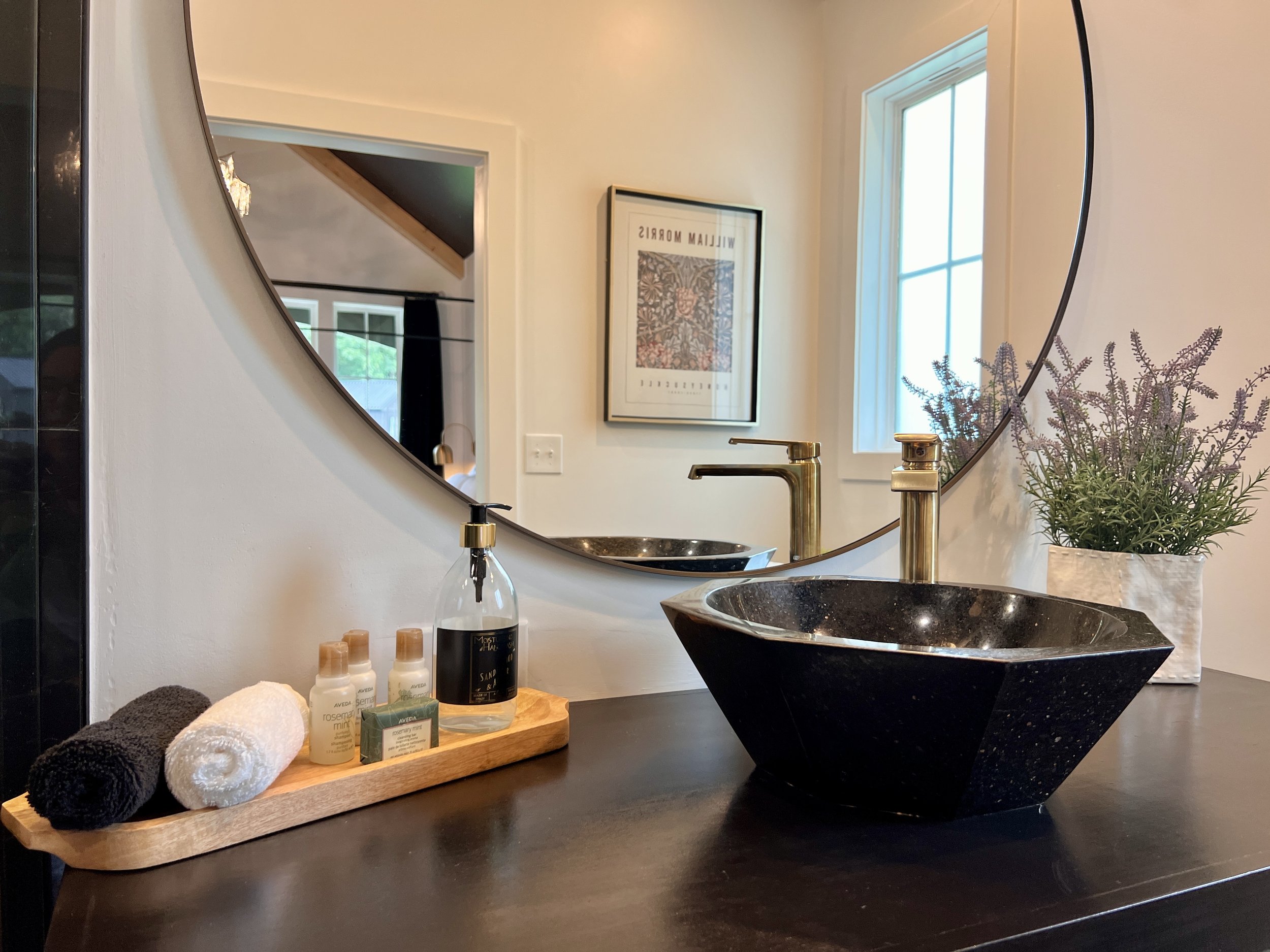 Modern bathroom sink area with black vessel sink and gold faucet, large round mirror, wooden tray with rolled towels and toiletries, and potted lavender plant.