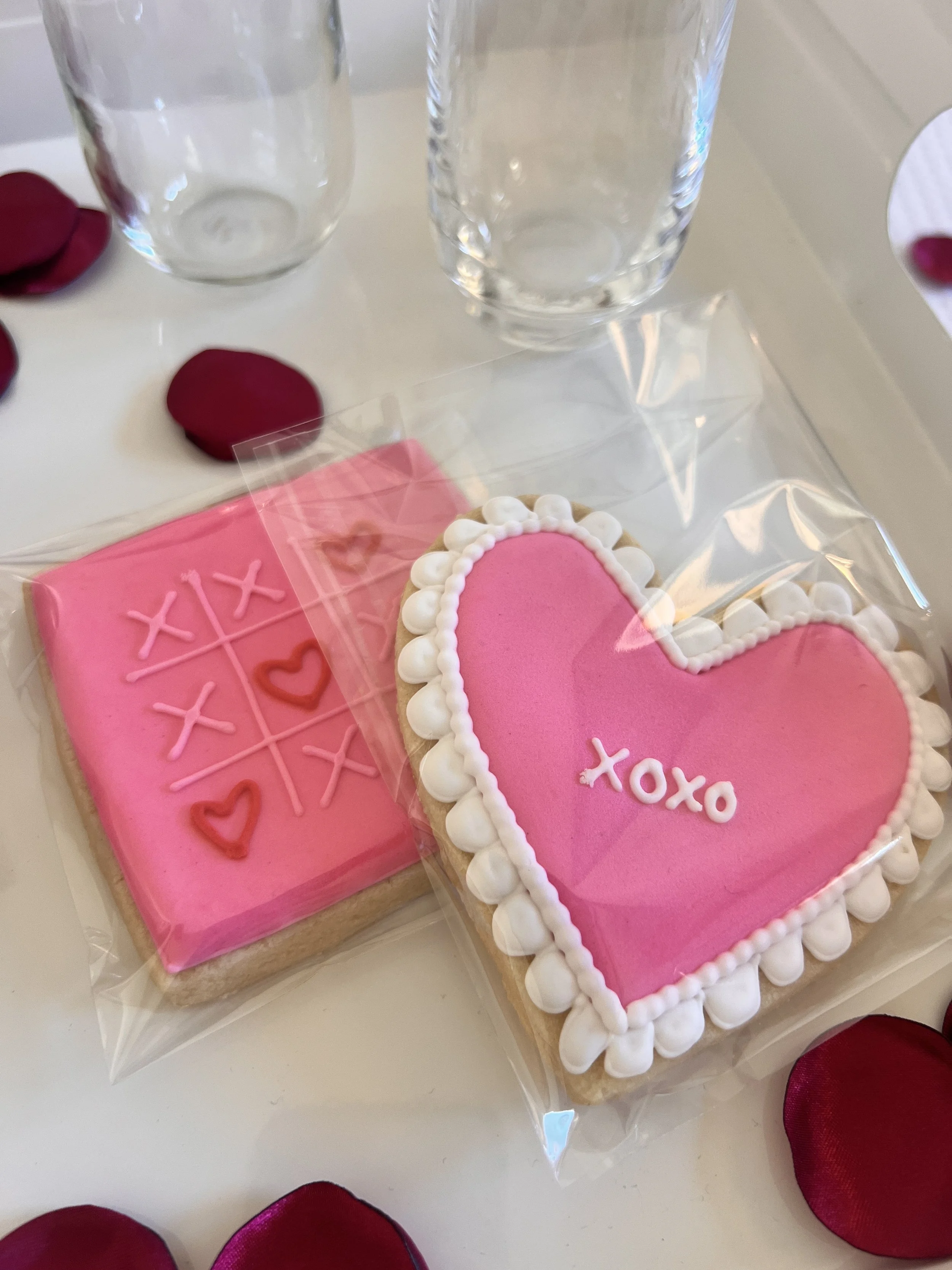 Two pink cookies packaged in clear bags, surrounded by rose petals. One cookie is square with a tic-tac-toe design and hearts, the other is heart-shaped with "xoxo" writing and white frosting accents. Glasses are visible in the background.