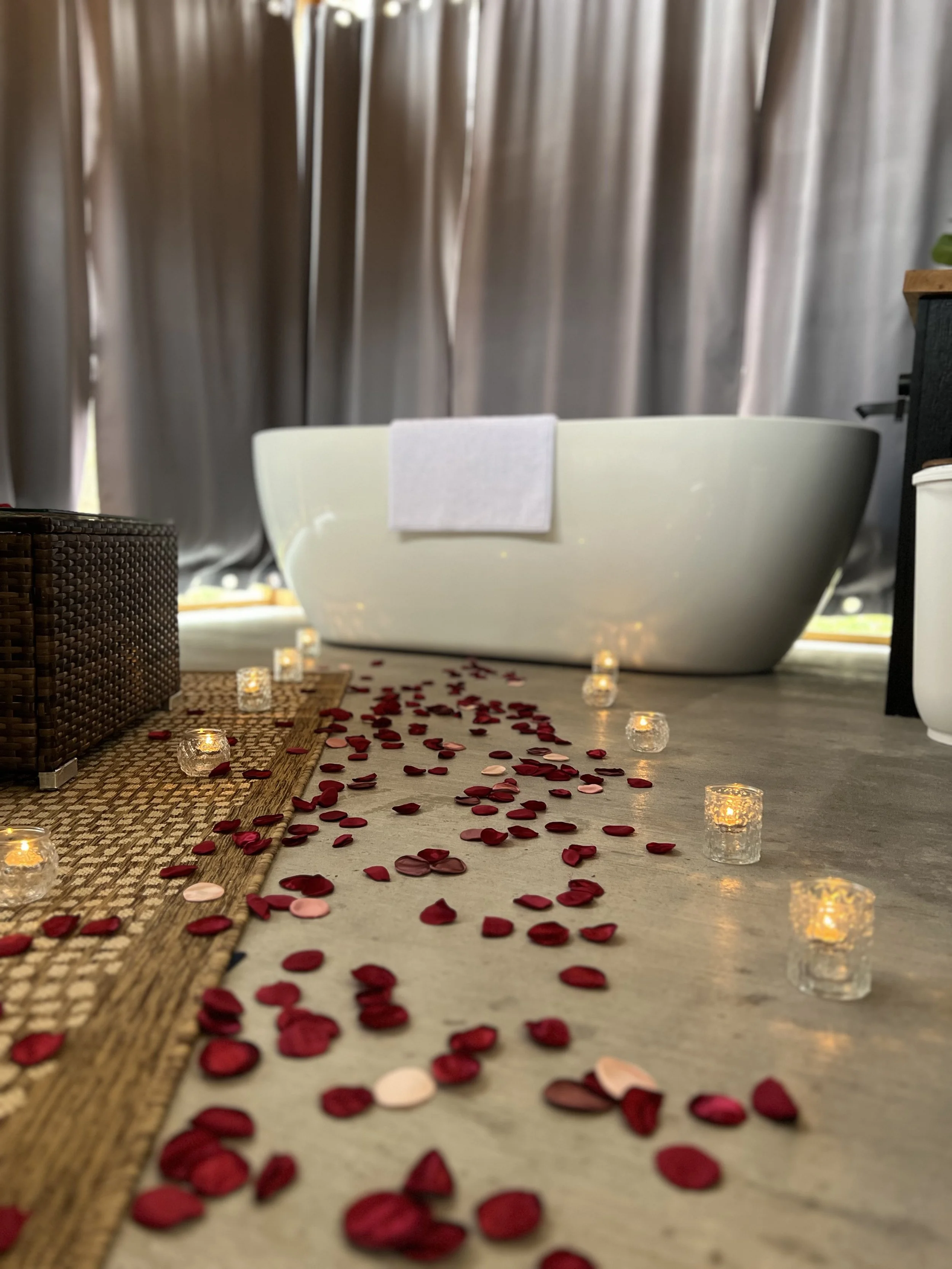 Bathtub with a towel, surrounded by rose petals and lit candles, in a luxurious setting with curtains.