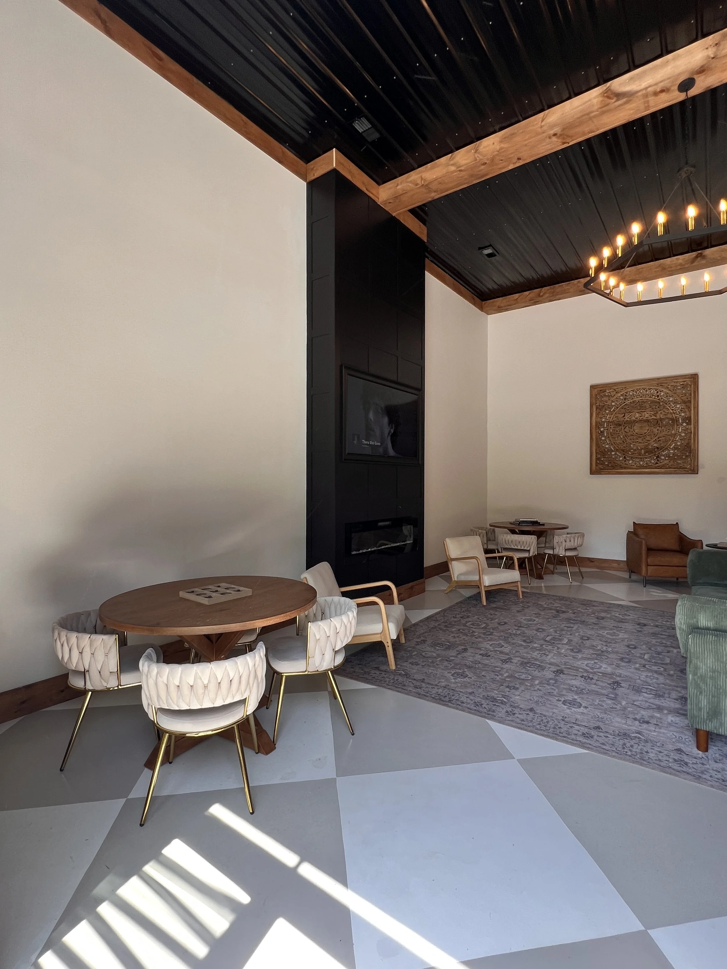 Modern lounge area with stylish furniture, including a round wooden table surrounded by upholstered chairs, a fireplace, and a large decorative ceiling light. The room has geometric tiled floors and a patterned rug, with a mix of light-colored and wo