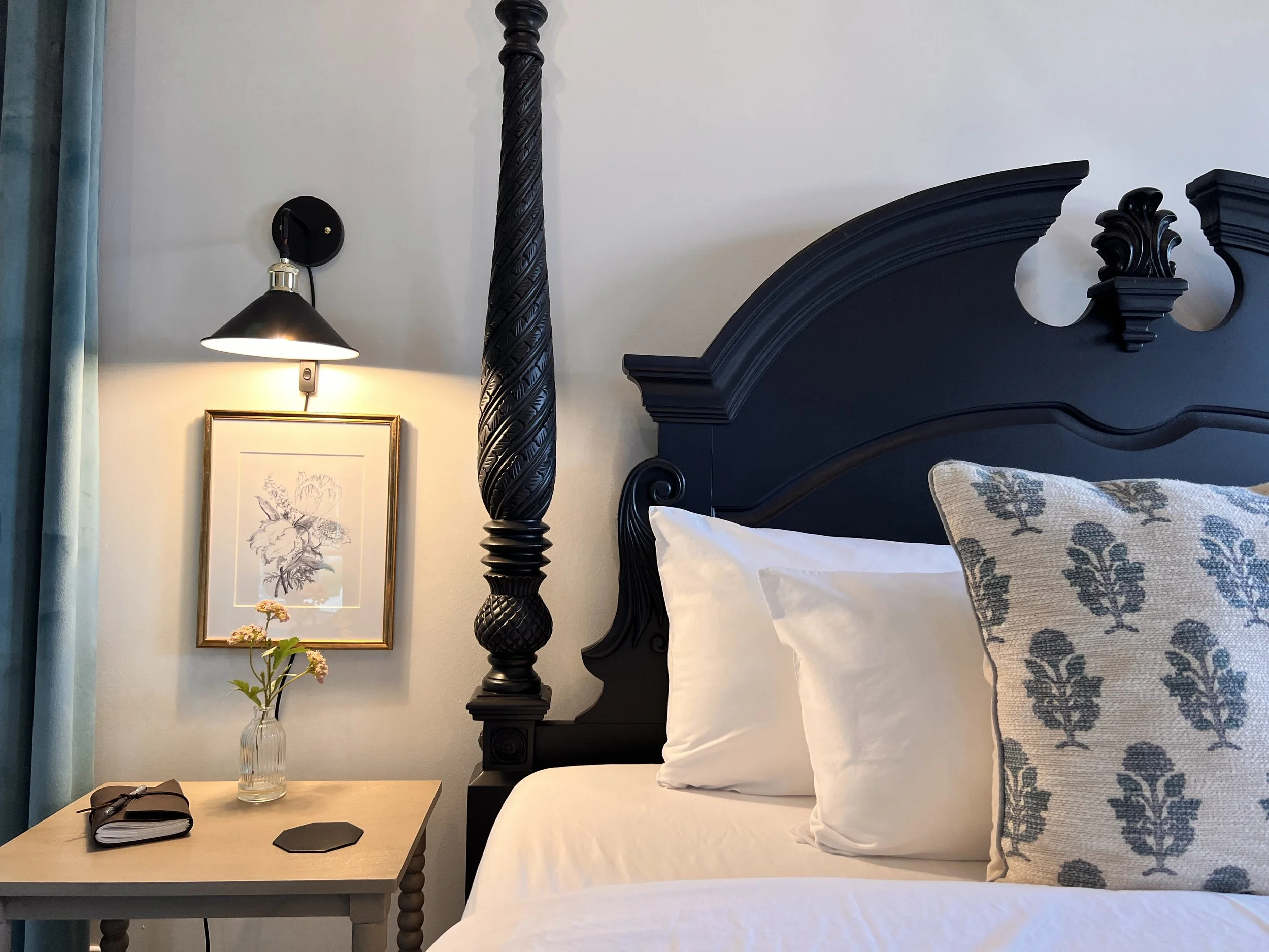 Cozy bedroom setting with a dark wooden bedframe, decorative pillows, a bedside table with a vase of flowers, a book, and a wall-mounted light above framed artwork.