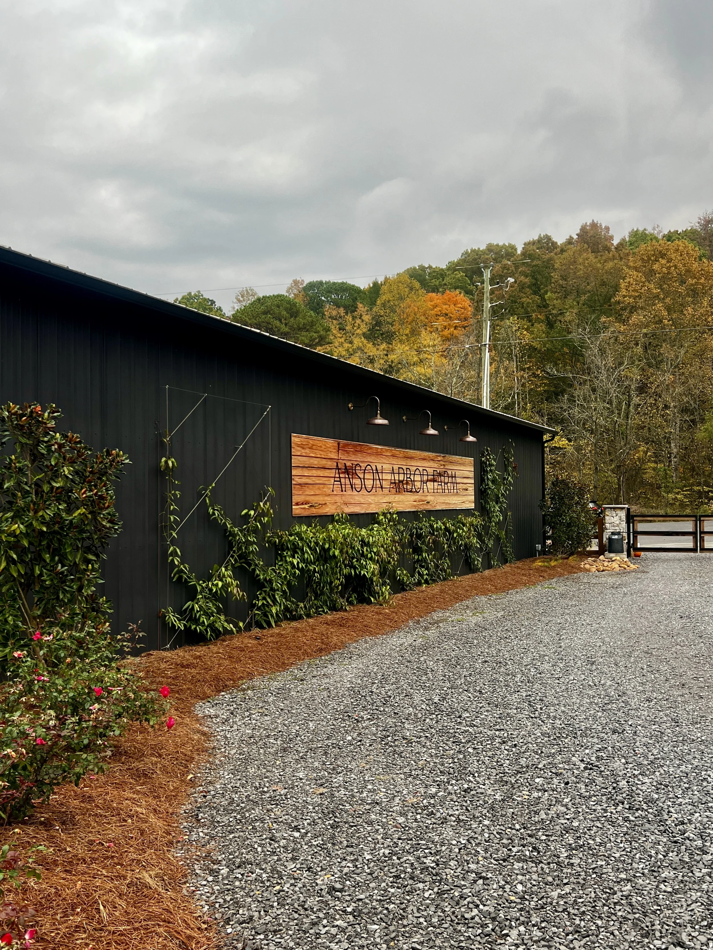 Exterior of a black building with a wooden sign reading "Anson Arbor Farm." The foreground has a gravel path, and the background features trees with autumn foliage under a cloudy sky.
