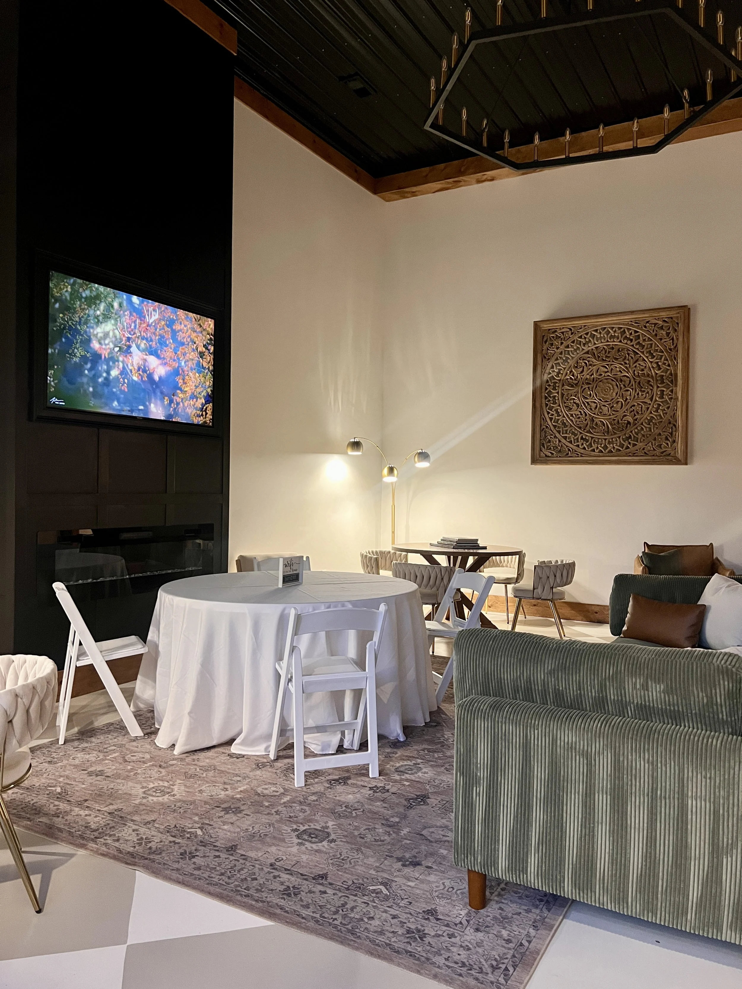 Elegant lounge area with a round table covered in a white tablecloth, surrounded by white chairs. A patterned rug underneath. A flat-screen TV on the dark wall and a decorative wooden panel next to it. Green upholstered seating and modern lighting co