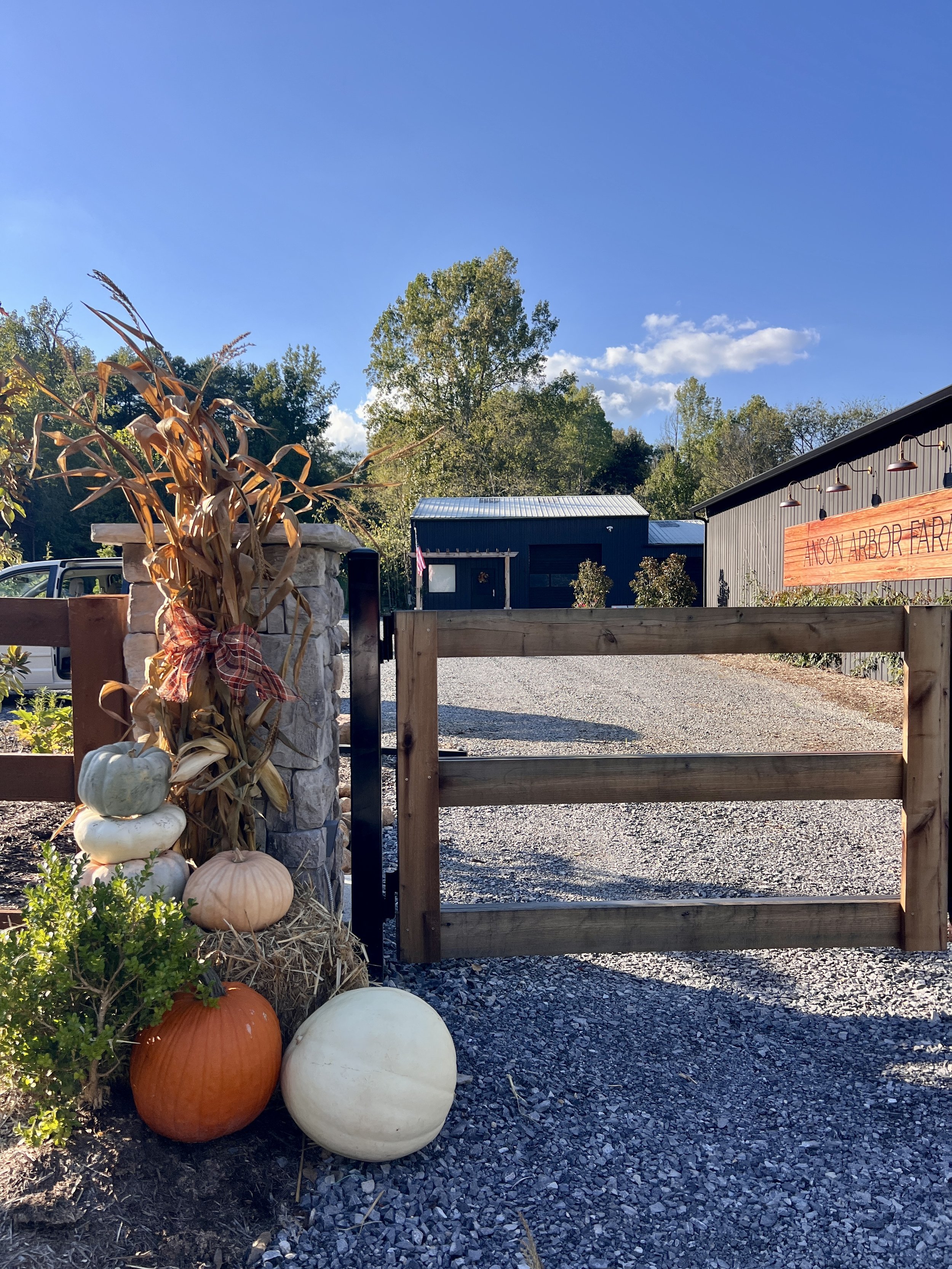 Entrance to a farm with a wooden gate, decorated with pumpkins and corn stalks. A sign reads 'NECK ARBOR FARM' on a barn. Trees and clear sky in the background.