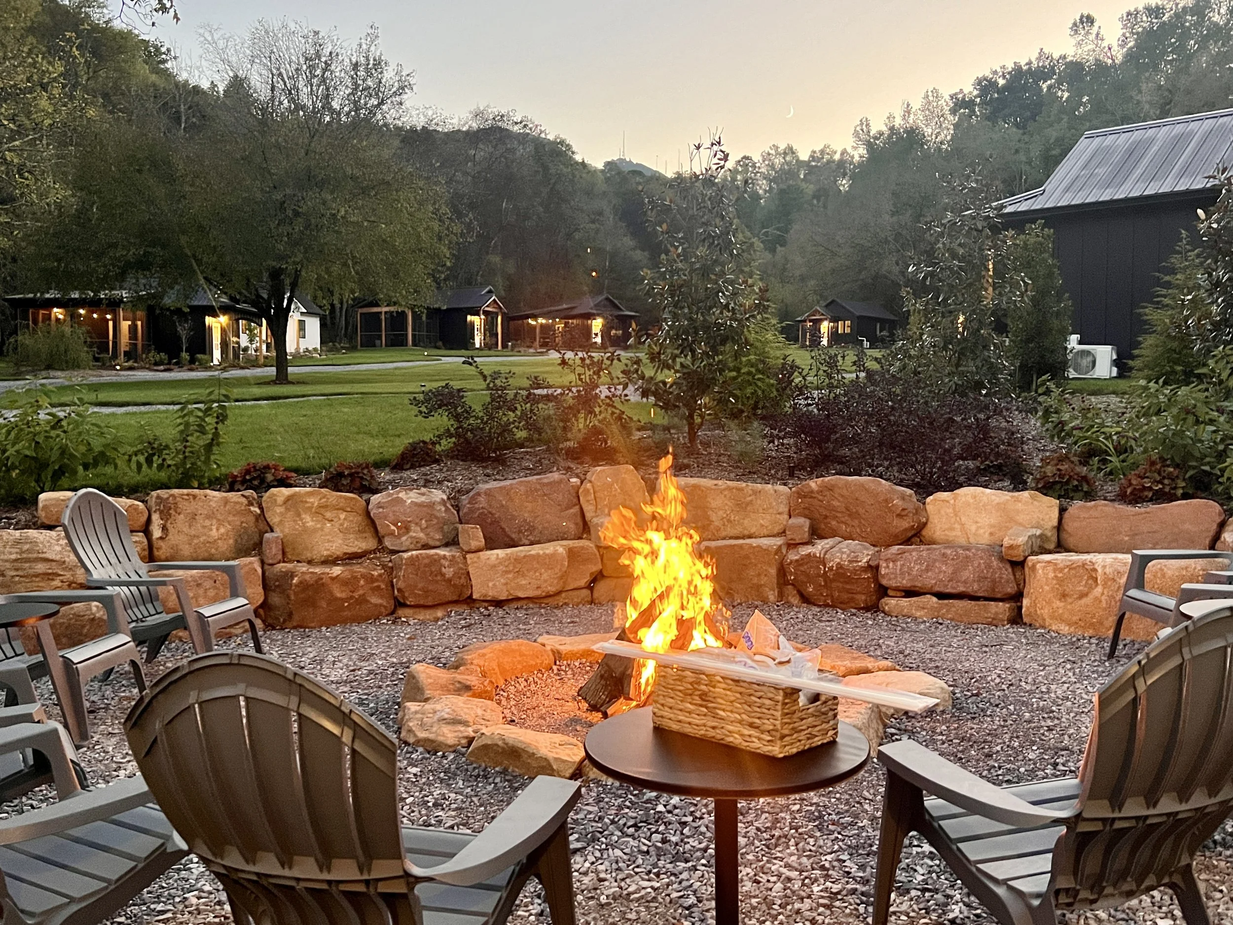 Outdoor fire pit with chairs surrounded by trees and buildings in a woodsy setting at dusk.
