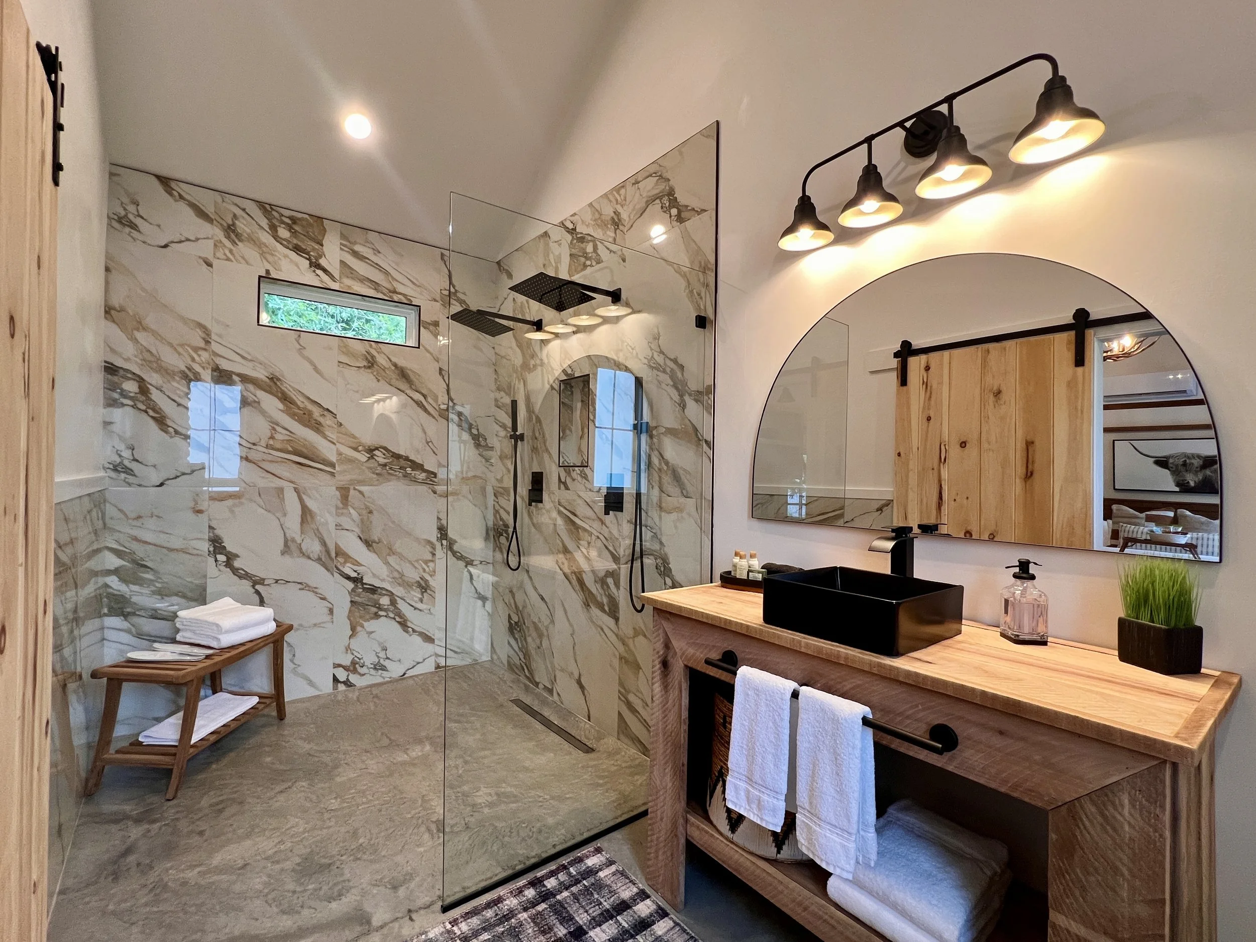 Modern bathroom with marble-tiled walk-in shower, wooden vanity with black sink, and round mirror. Minimalist decor with towels and small plant.