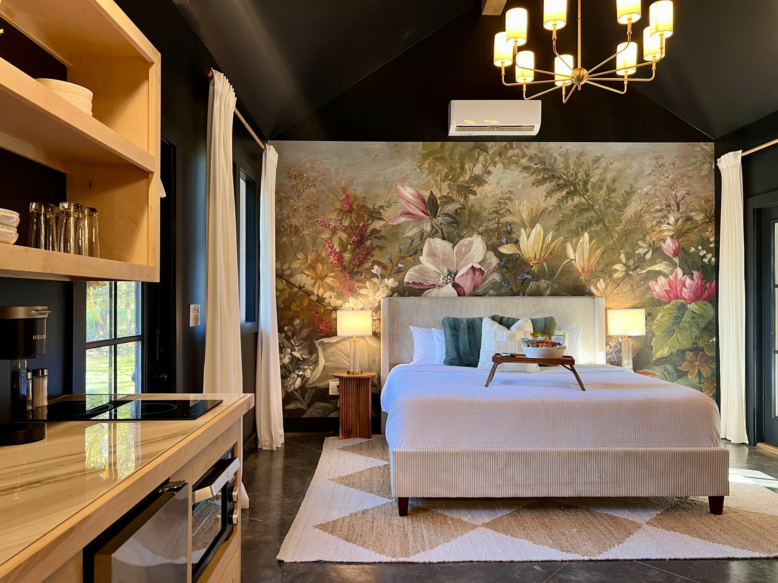 Luxurious bedroom with floral mural, modern chandelier, and kitchenette featuring coffee maker and toaster oven.