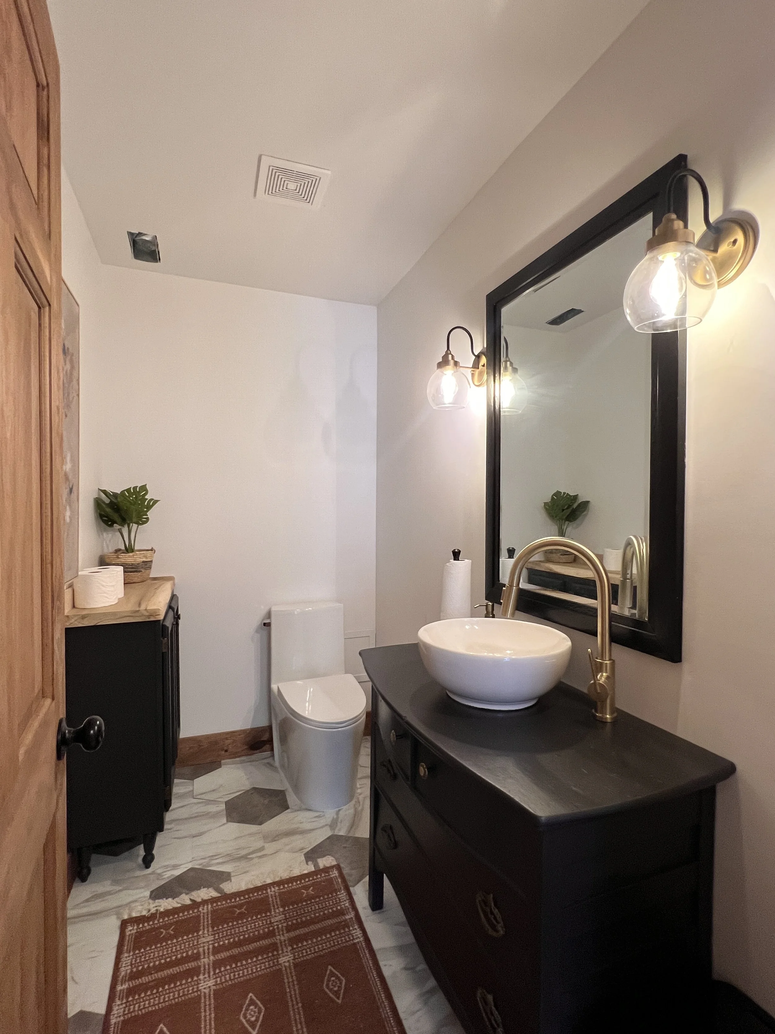 A modern bathroom with a black vanity featuring a round white vessel sink and a brass faucet. A large mirror above the sink is flanked by two wall sconces with clear glass globe shades. A toilet is visible in the background next to a small cabinet wi