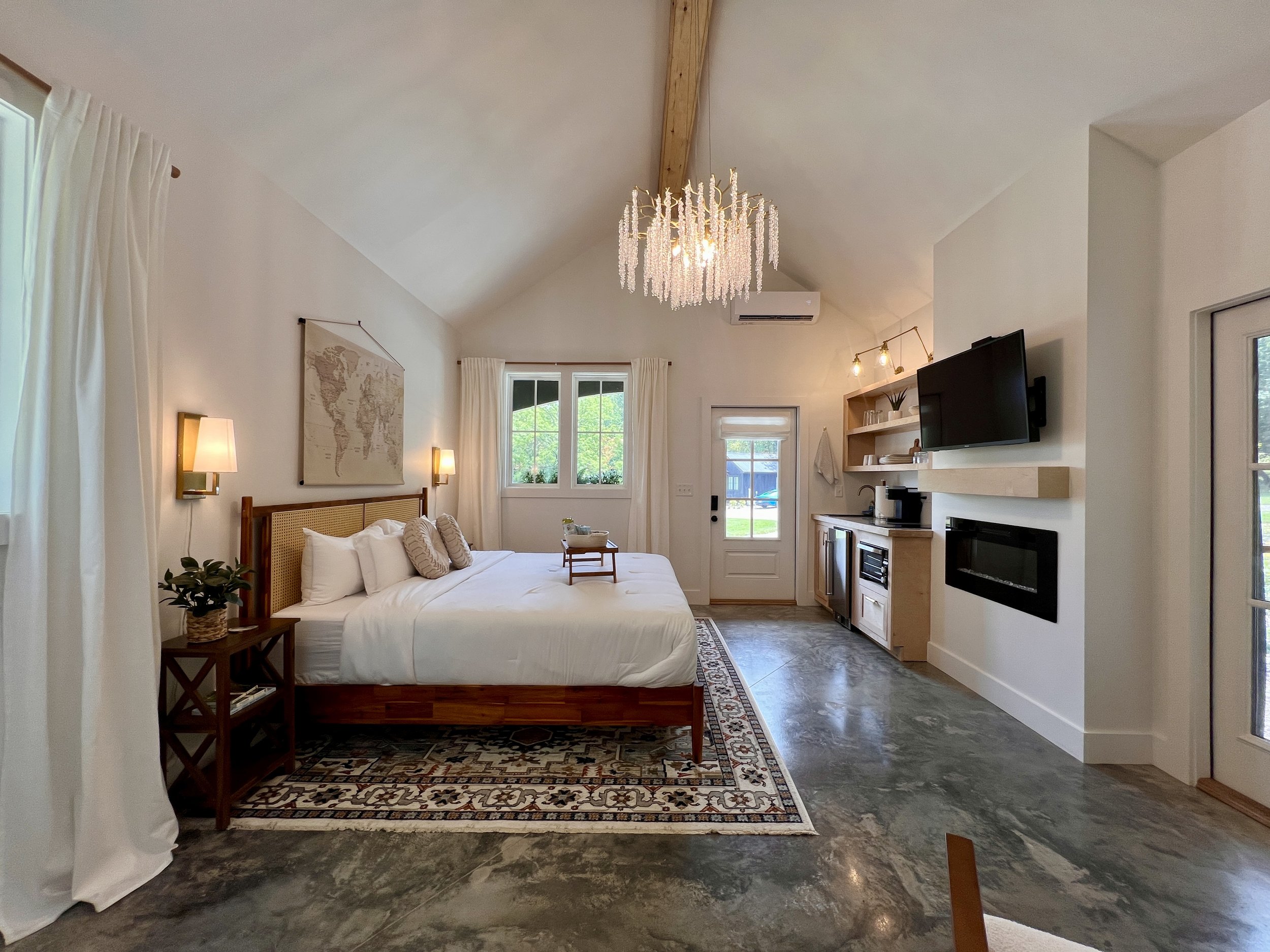 Modern bedroom with a king-sized bed, chandelier, large map artwork, decorative rug, wall-mounted TV, fireplace, kitchenette, and large windows. The room has a high ceiling and polished concrete floor.