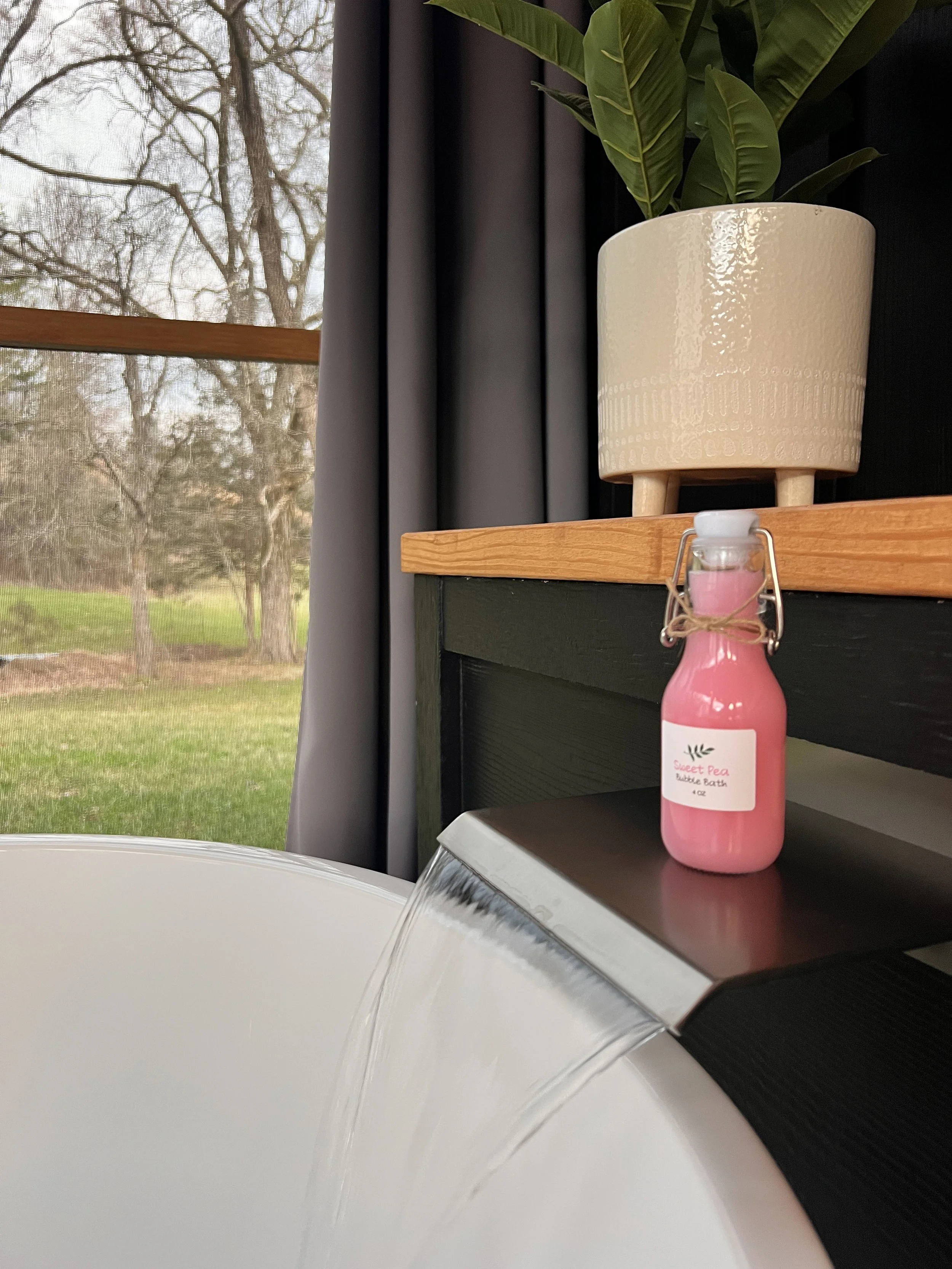 A modern bathroom with a white freestanding bathtub filled with water, a small pink bottle labeled "Sweet Pea Bubble Bath," and a potted plant in a decorative pot. A large window in the background displays an outdoor view of trees and grass.