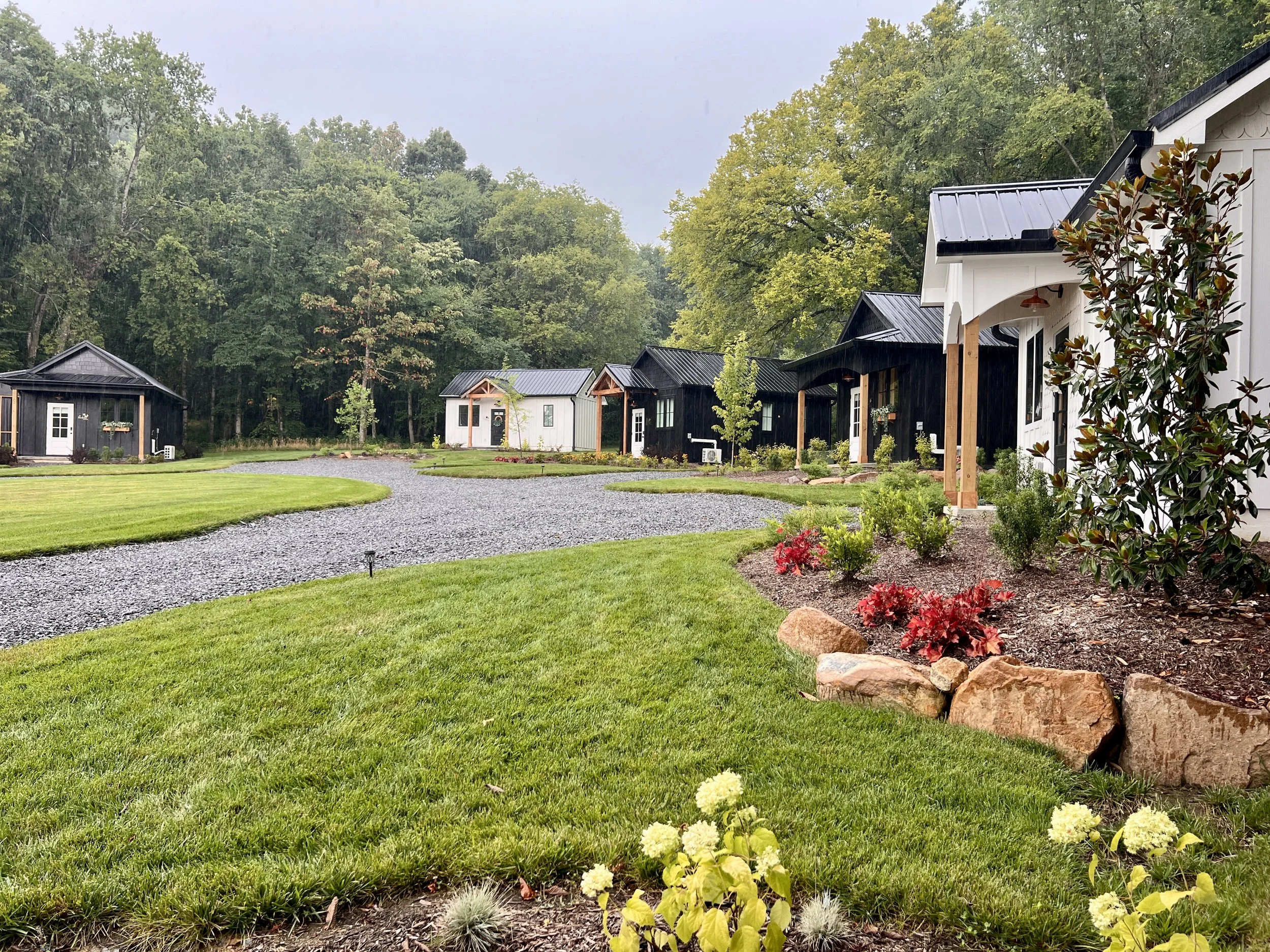 A serene outdoor setting with small black and white cottages surrounded by lush green grass, gravel paths, and landscaped gardens. Trees and shrubs provide a natural backdrop, enhancing the peaceful ambiance.