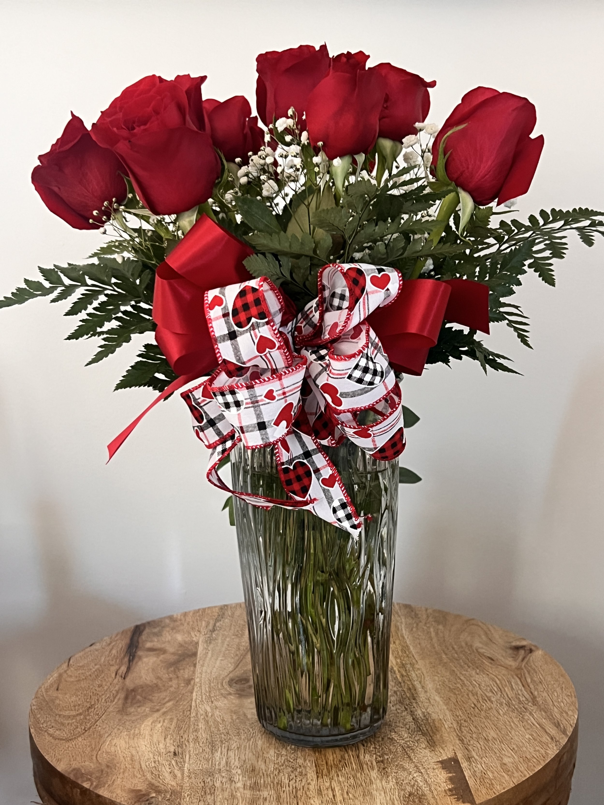 Bouquet of red roses in a clear glass vase with decorative ribbons on a wooden table.