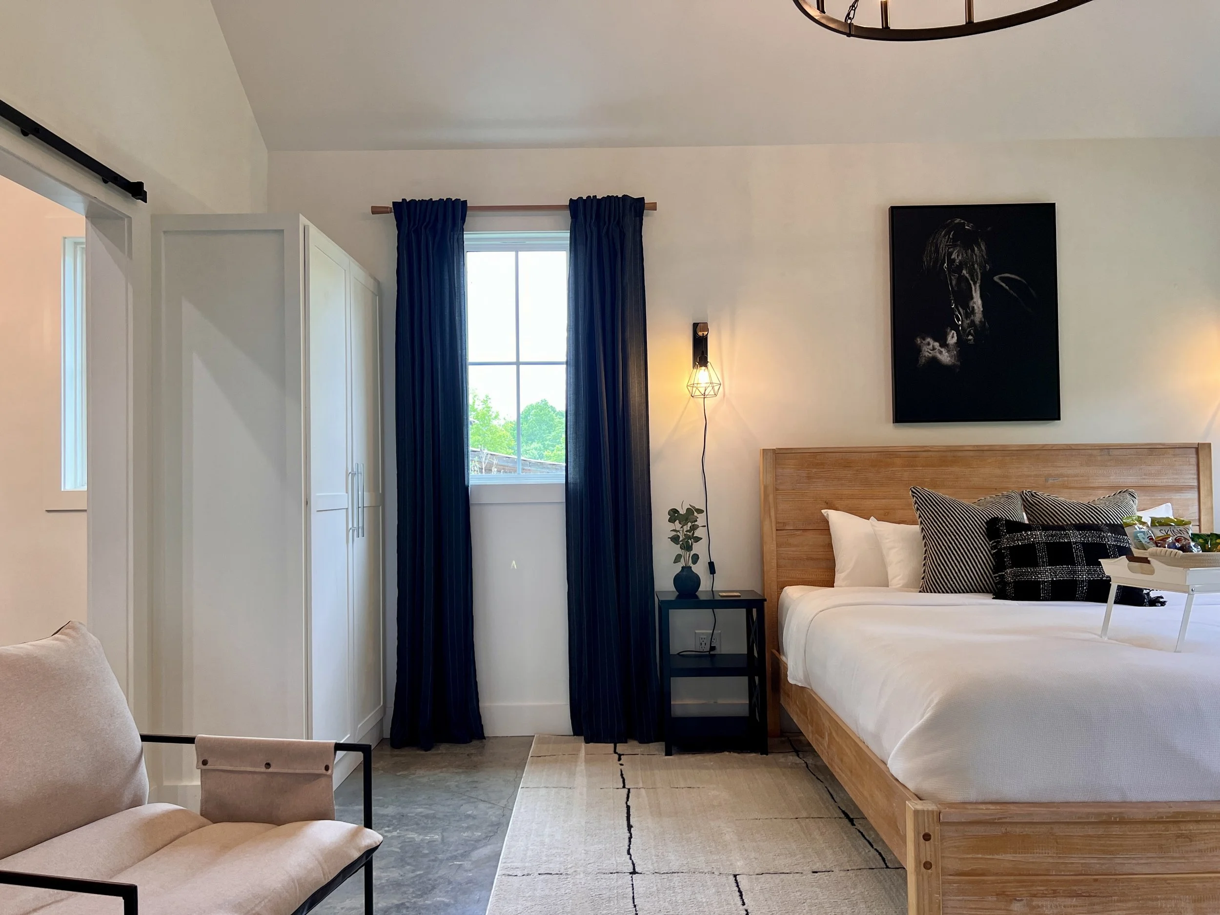 Modern bedroom interior with a wooden bed, white bedding, black pillows, and a side table with a vase. There's a piece of wall art above the bed, a large window with blue curtains, a white wardrobe, and a beige chair.