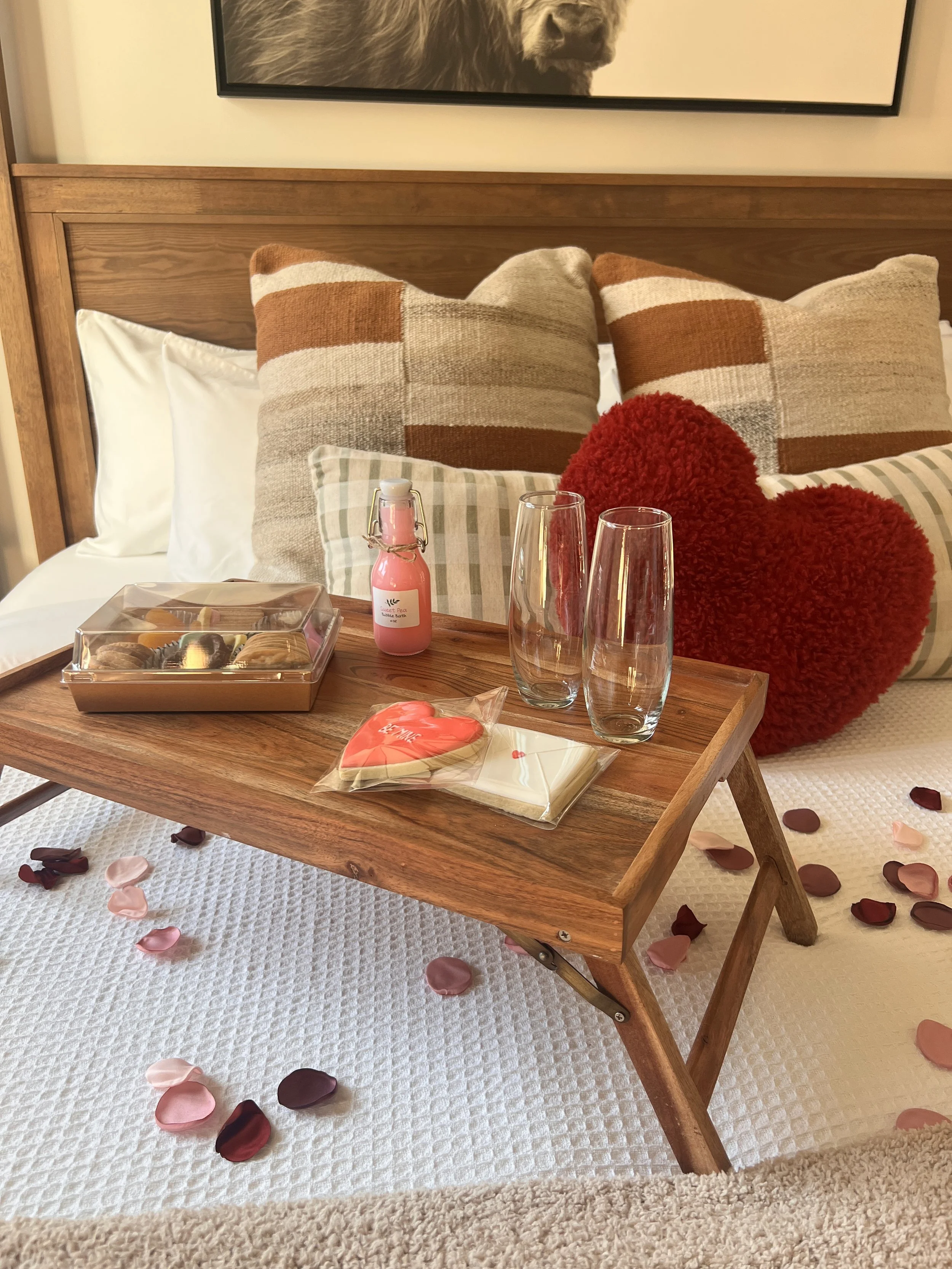 Bed with a wooden tray holding a pink drink, heart-shaped cookie, chocolates, and two glasses, decorated with heart and striped pillows and rose petals.