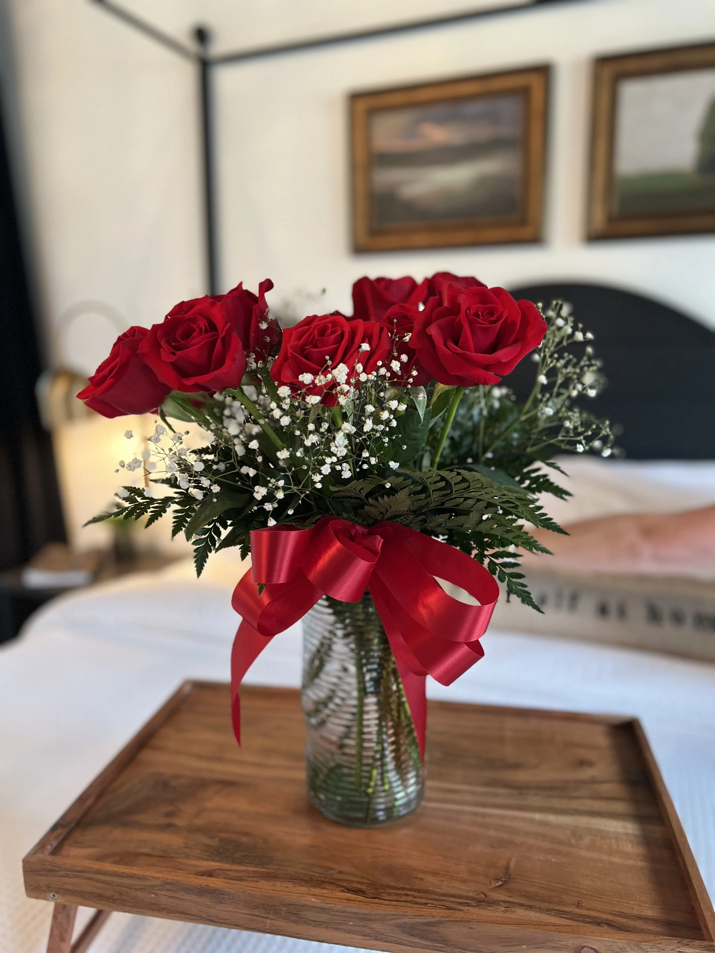 Bouquet of red roses with baby's breath and fern foliage in a glass vase, tied with a red ribbon, on a wooden tray.