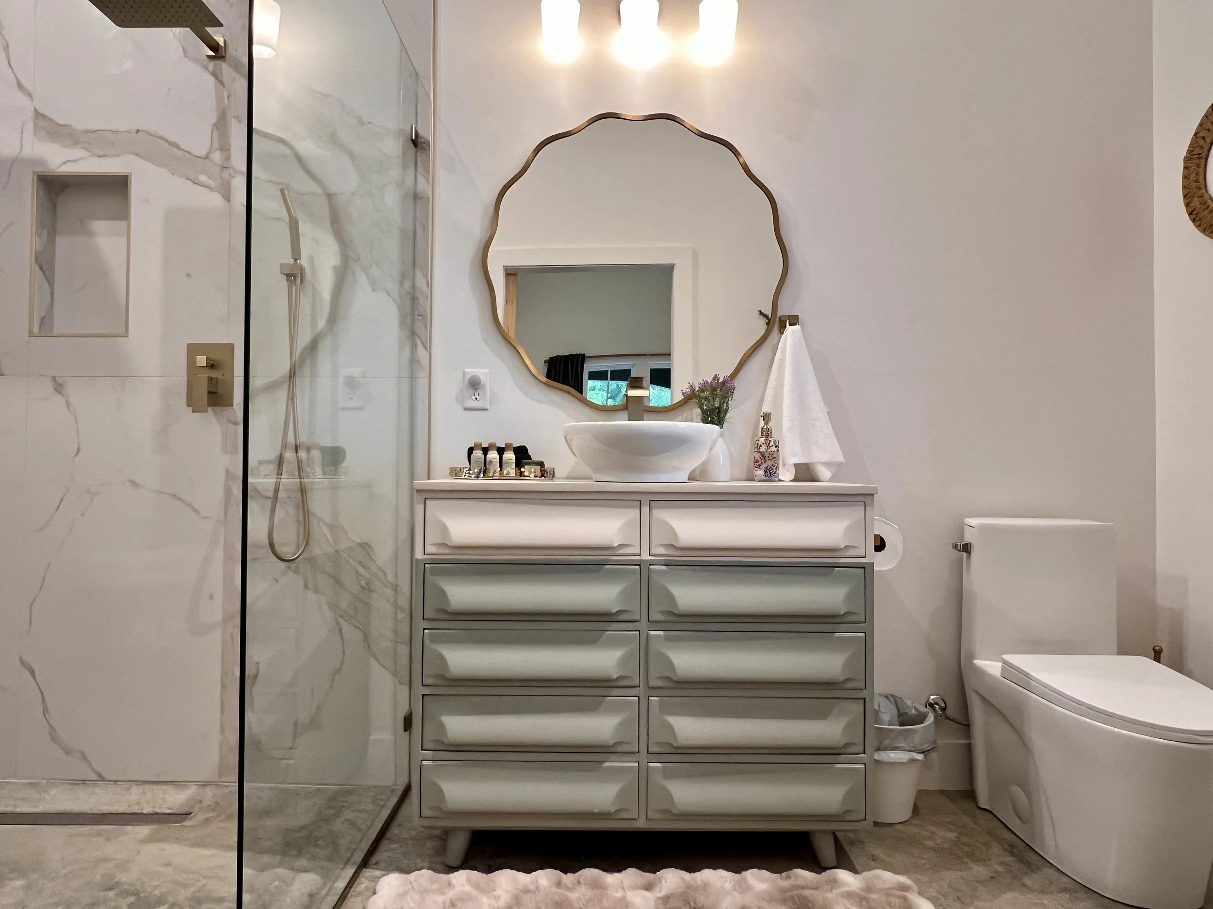 Modern bathroom with a glass shower, marble walls, floating vanity, vessel sink, mirror with decorative frame, and contemporary toilet.