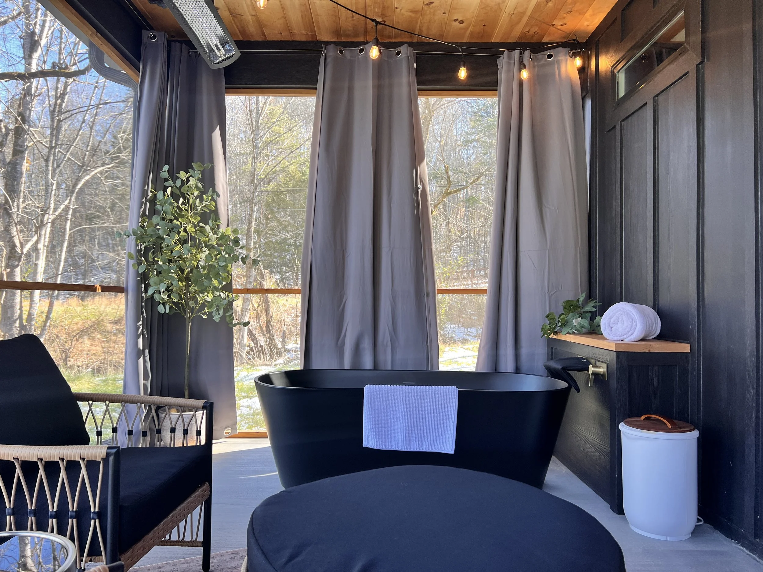 Outdoor bath area with a black bathtub, gray curtains, and cozy seating. Surrounded by nature, featuring a potted plant, rolled towel, and decorative lights.