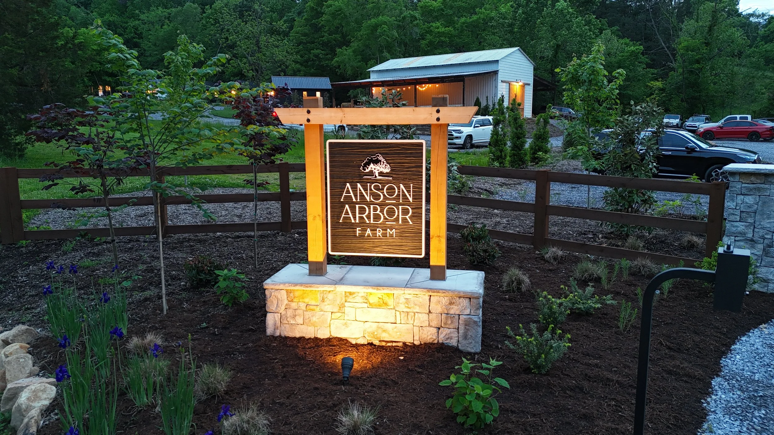 Entrance sign for Anson Arbor Farm surrounded by landscaping, a wooden fence, and a building in the background.