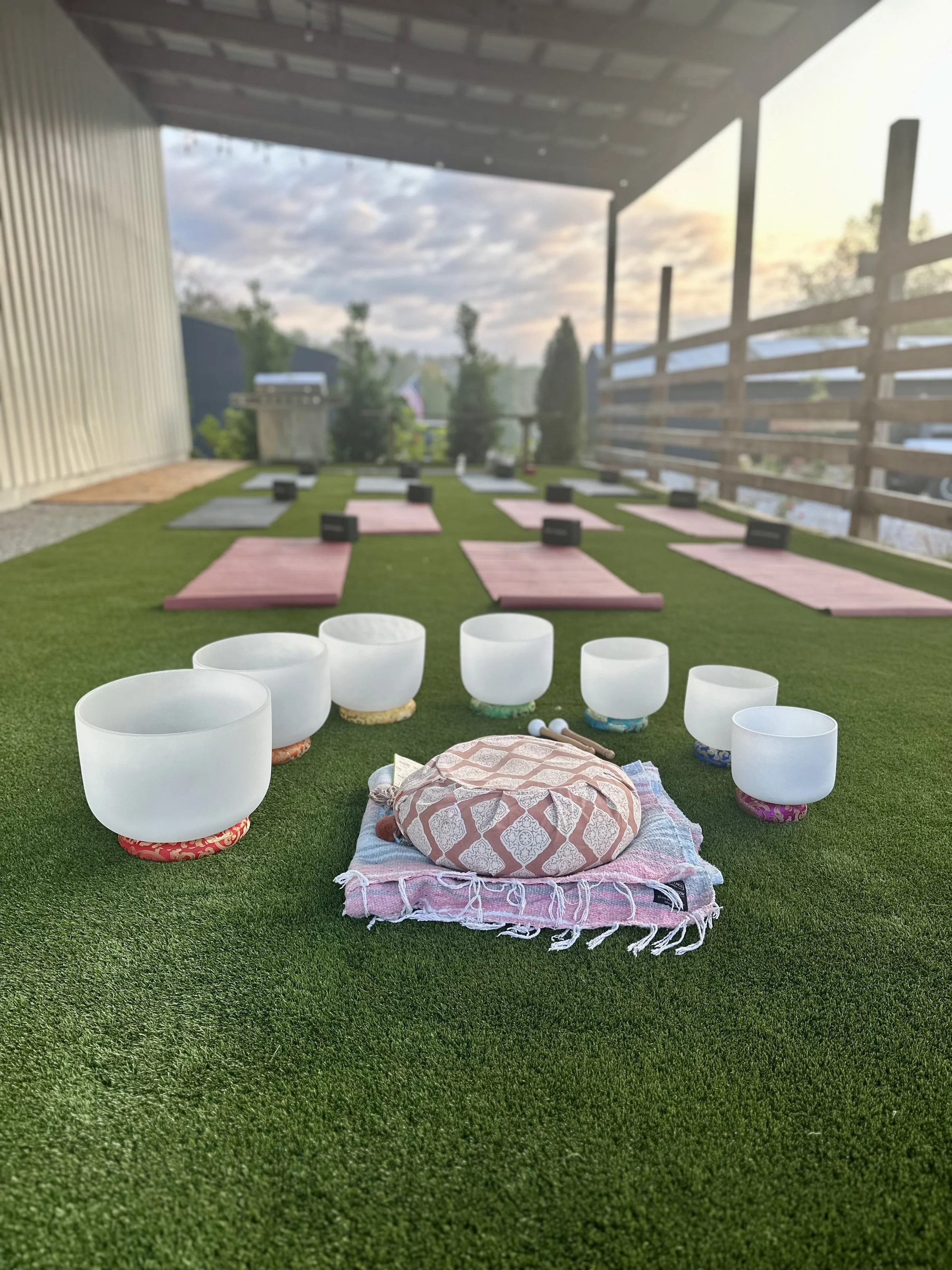 Outdoor yoga and meditation setup with singing bowls and cushions on grass, surrounded by yoga mats and blocks, under a covered patio with sunset view.