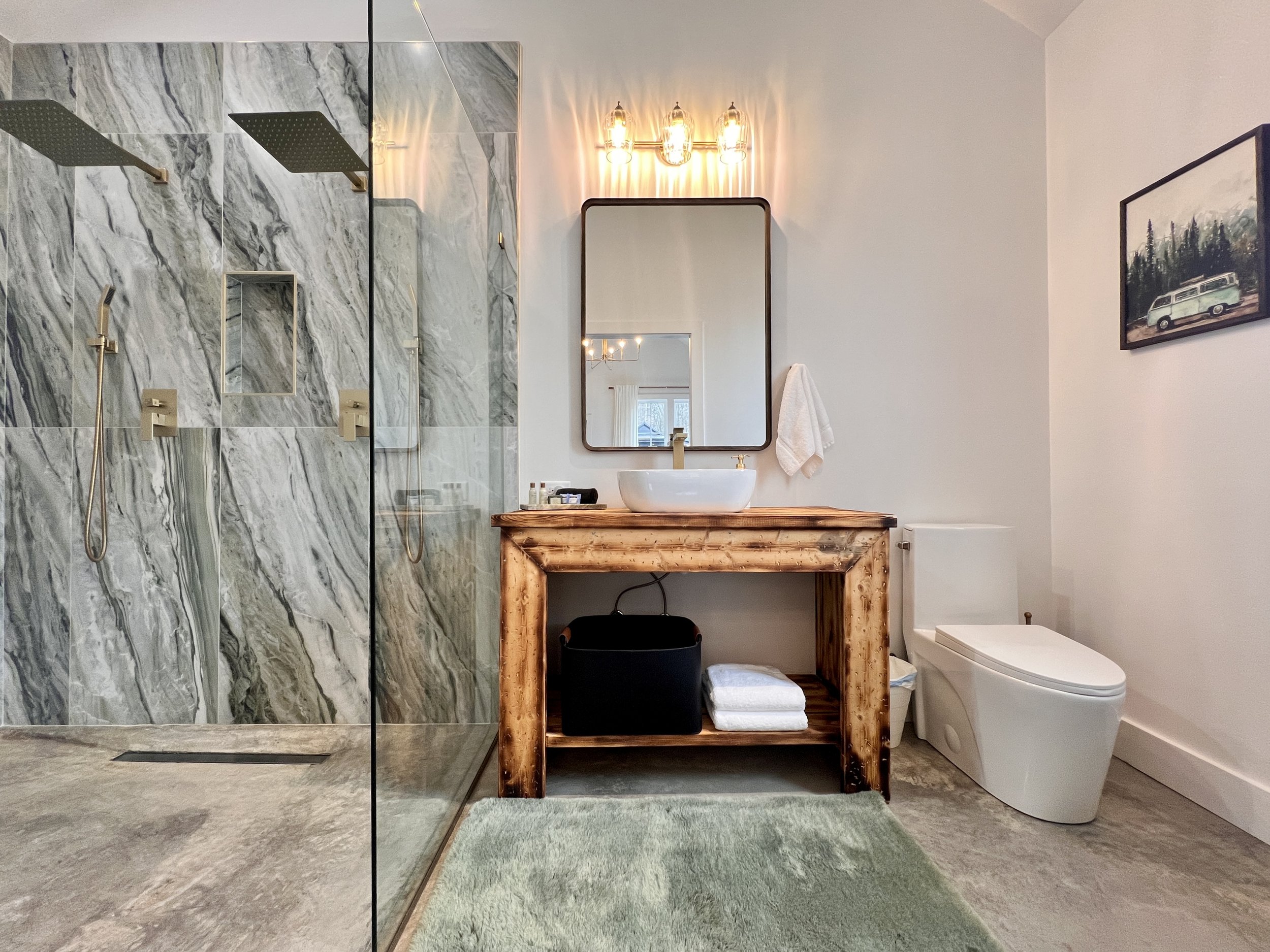 Modern bathroom with a large glass-enclosed shower featuring marble walls and dual rain showerheads. The rustic wooden vanity has a vessel sink and a rectangular mirror above. The room includes a toilet, wall art, and soft lighting.