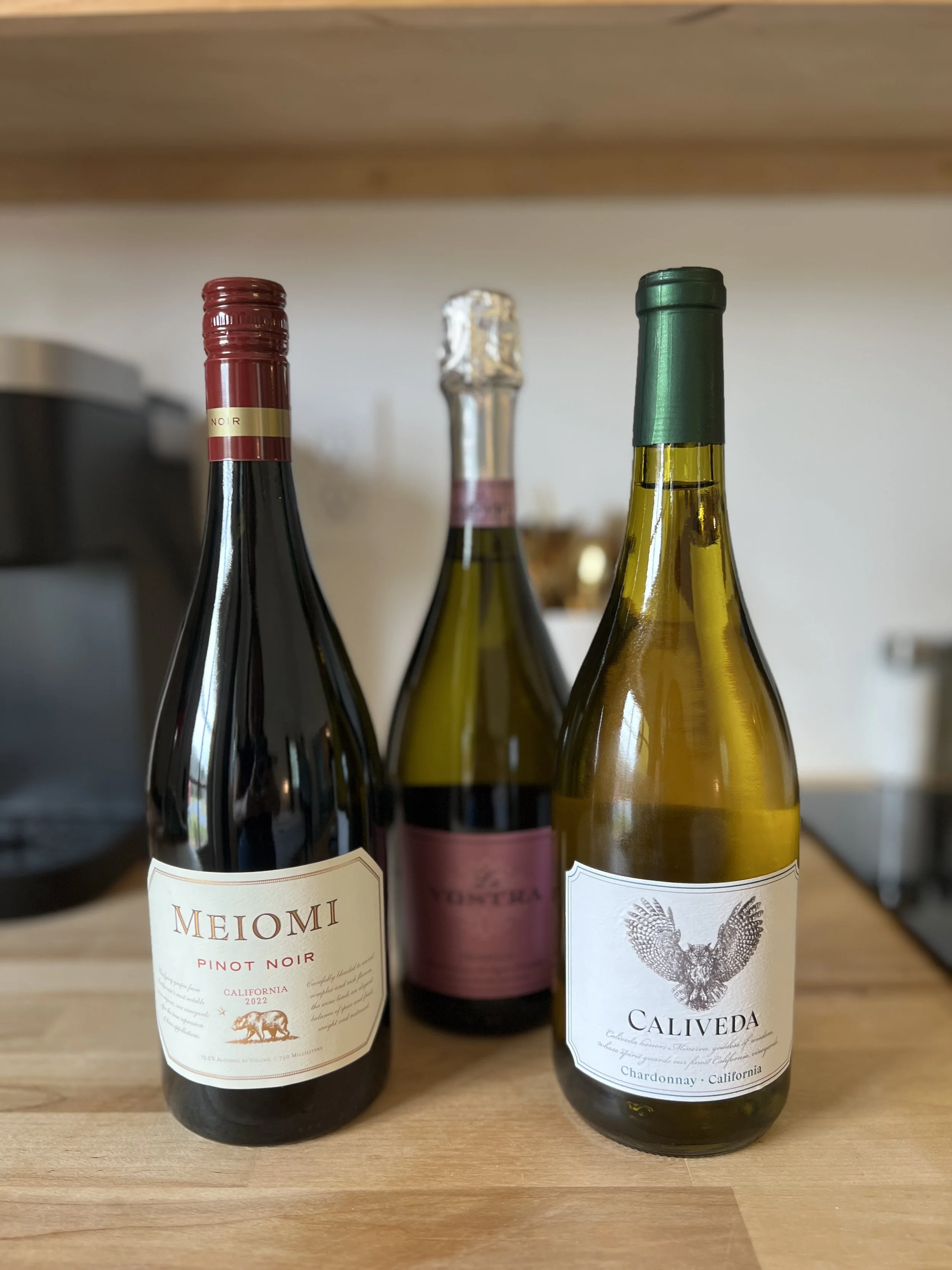 Three wine bottles on a wooden surface with a blurred background. The bottles include a red wine, sparkling wine, and a white wine. The first bottle on the left is a Pinot Noir, the middle bottle is a sparkling wine, and the bottle on the right is a 