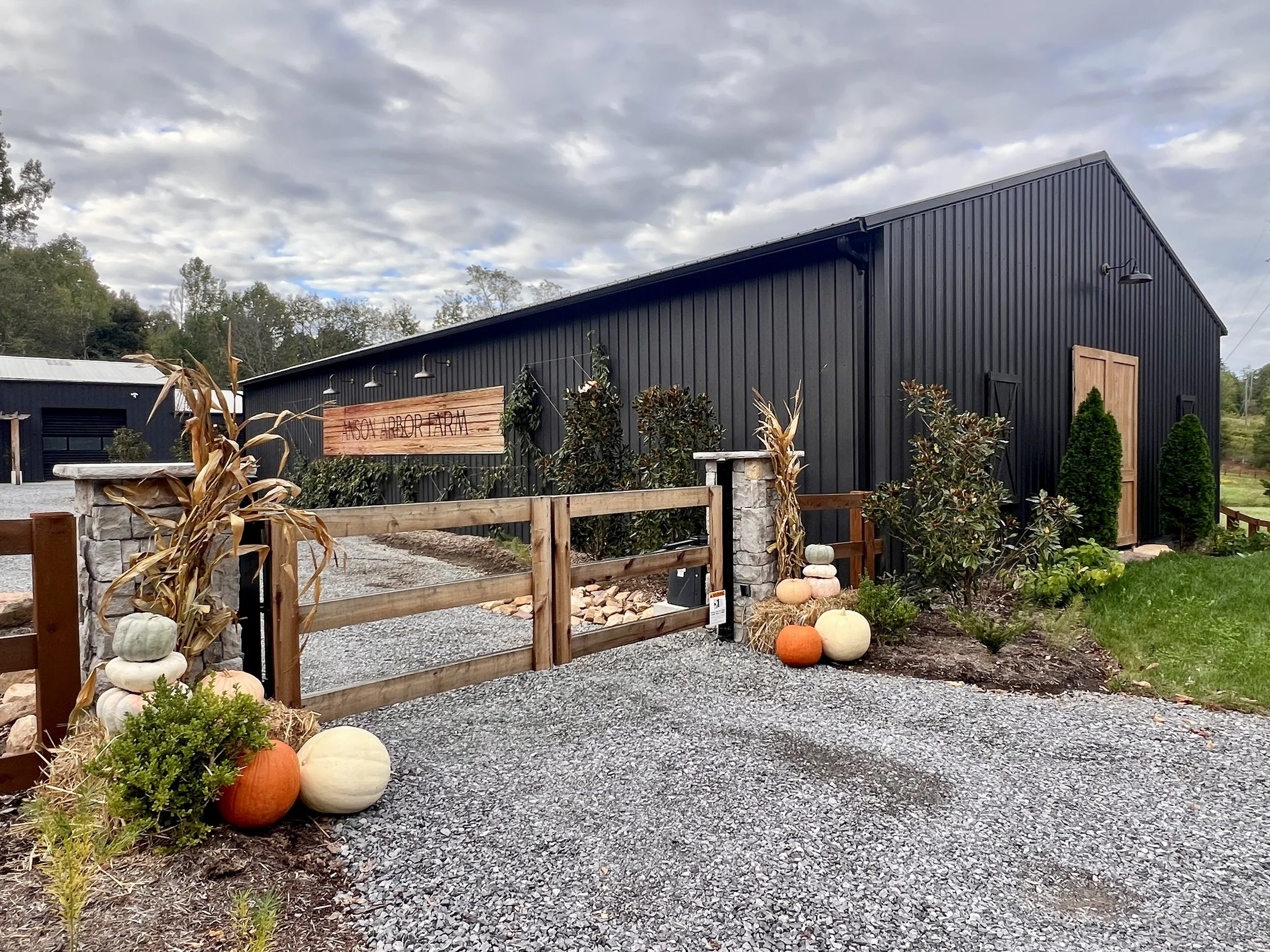 A rustic farm building with a sign reading "Noah Arbor Farm" near the entrance. The building is dark-colored with wooden doors, surrounded by gravel paths and greenery. Decorative pumpkins and corn stalks are placed on either side of the wooden gate.