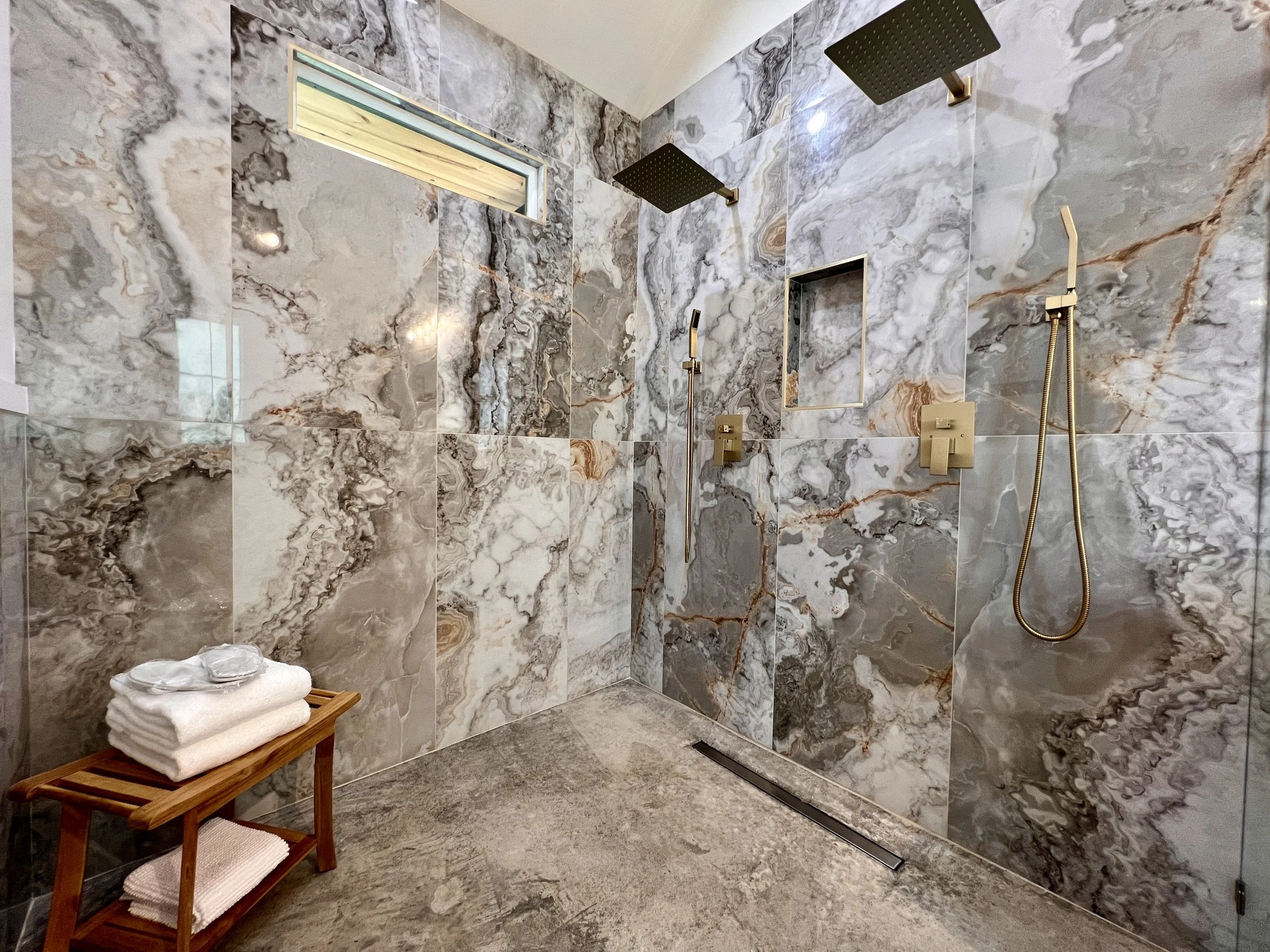 Luxurious shower with marble walls, two rain showerheads, a handheld shower, and a wooden bench with towels.