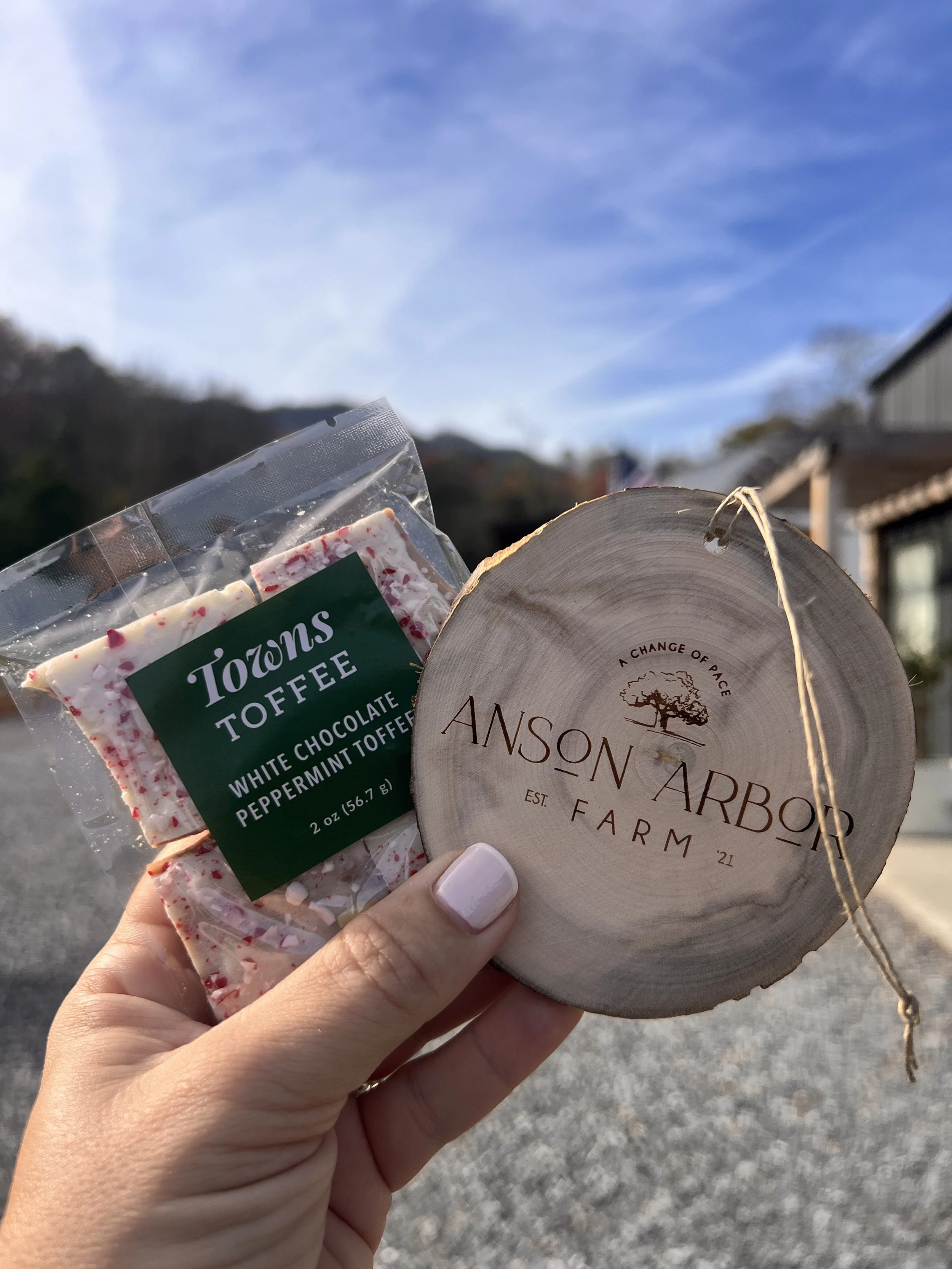 Close-up of a hand holding a package of Towns Toffee white chocolate peppermint toffee and a wooden tag from Anson Arbor Farm against a scenic outdoor background with blue sky.