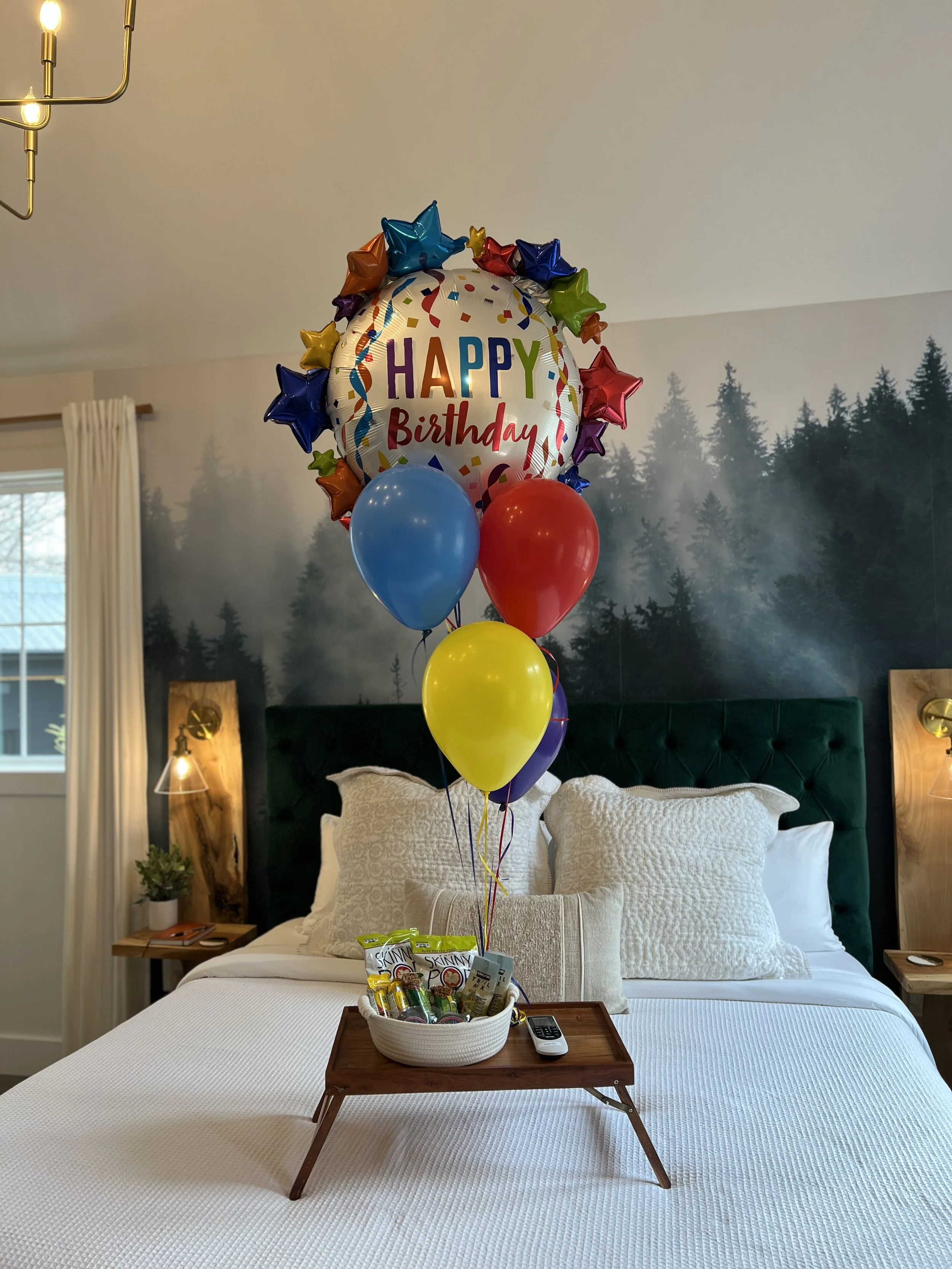 A bedroom with a mural of a forest and a bed with white linens. On the bed, there's a wooden tray holding a "Happy Birthday" balloon arrangement with colorful balloons and a basket of snacks. A remote control is beside the basket.