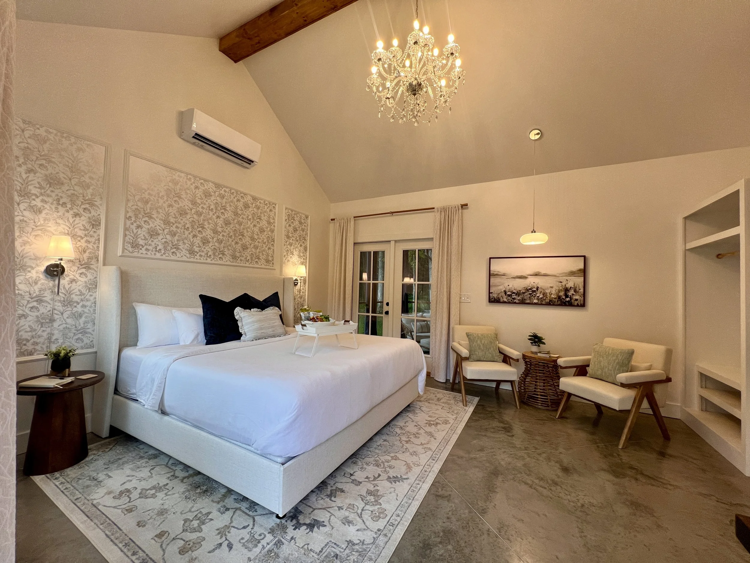 Elegant bedroom with a large bed, two armchairs, a chandelier, and a floral-patterned accent wall. The room includes a small round side table, gray area rug, framed artwork, and French doors.