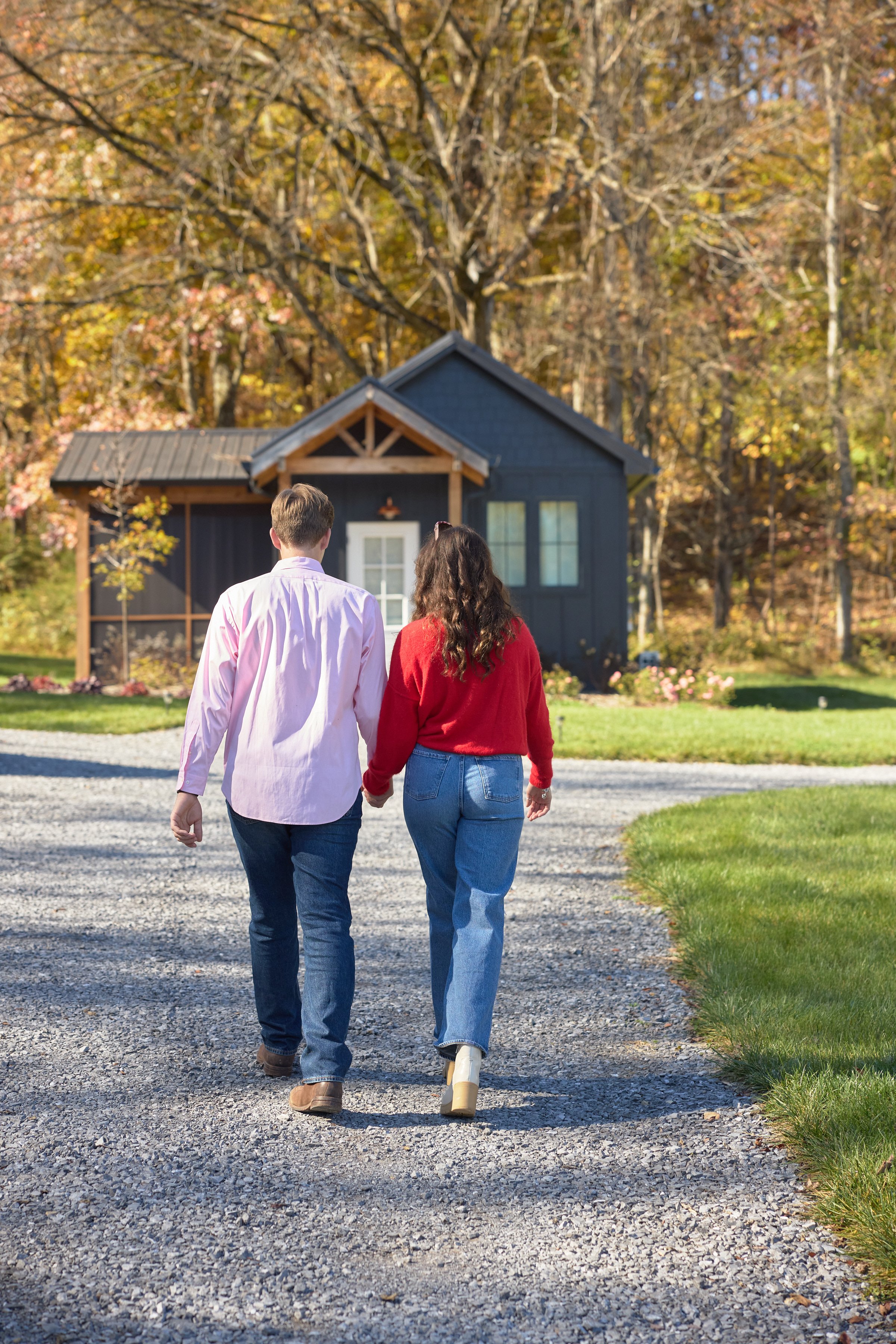 Couple walking hand in hand toward a small blue house surrounded by trees with autumn foliage.